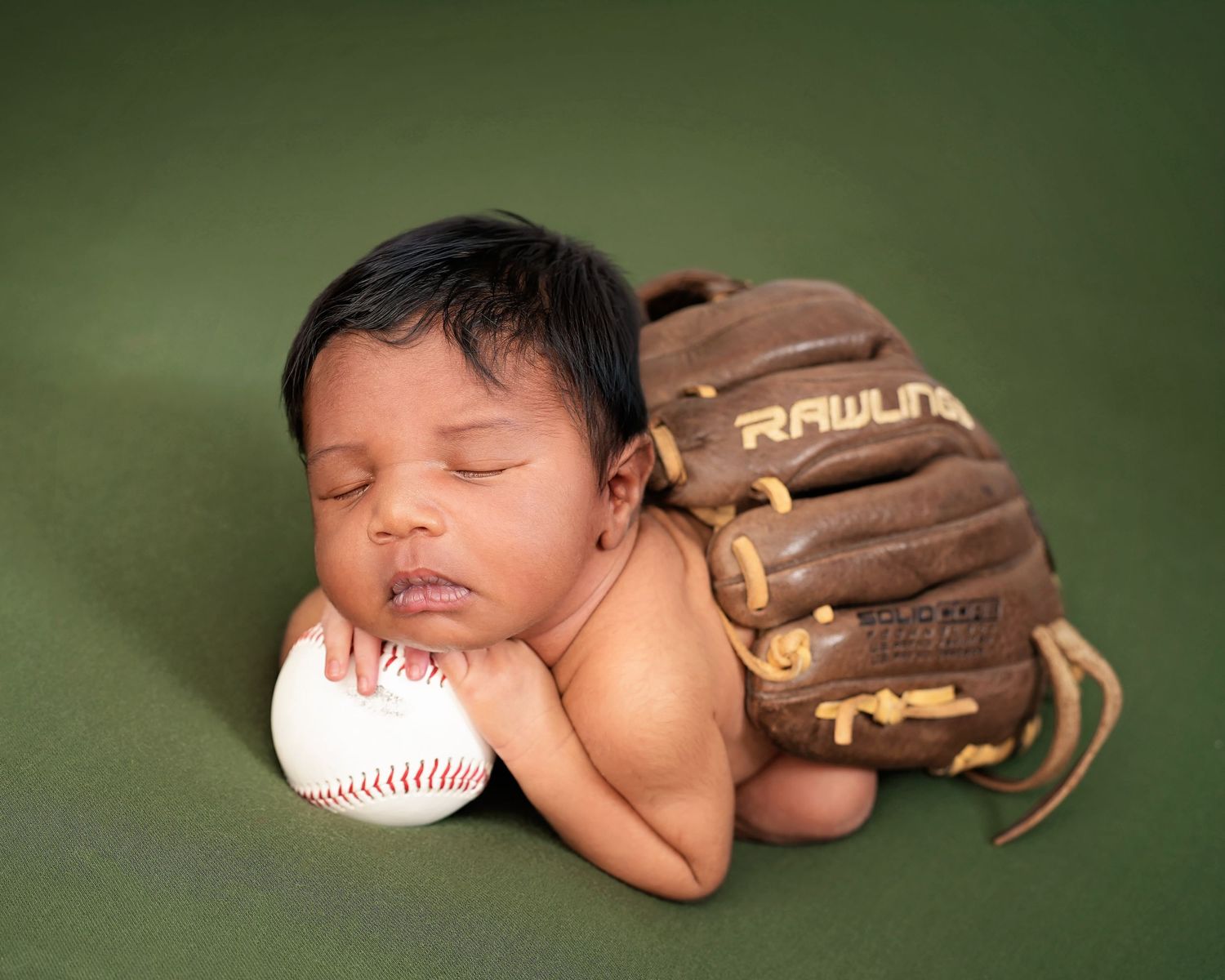 Newborn baby sleeping on a baseball with a Rawlings mitt on a green background.