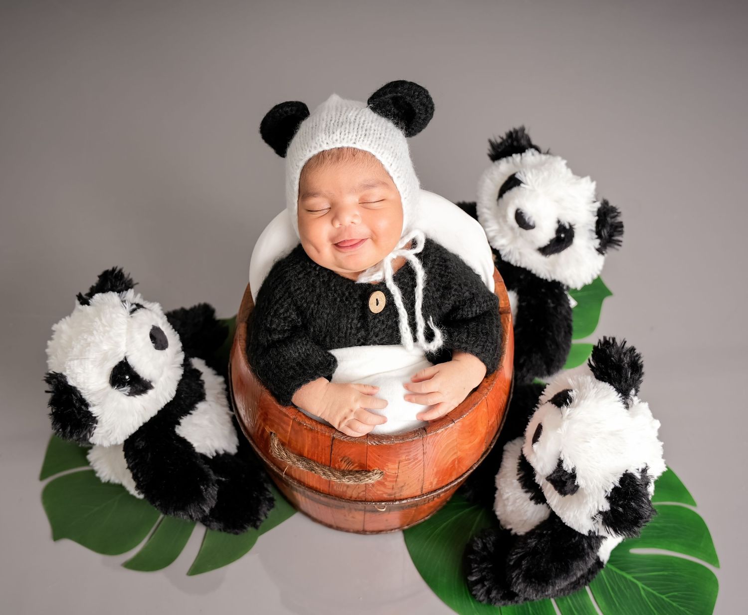 Baby in cozy panda hat sits in wooden basket surrounded by plush panda toys and green leaves on gray background.