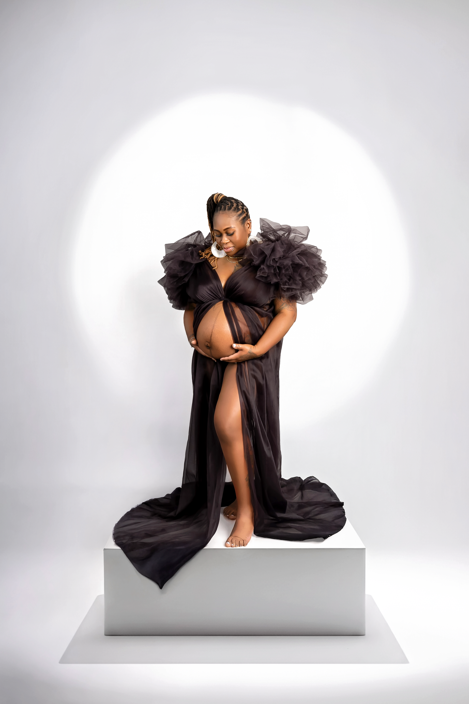 Artistic fashion portrait of a model in black ruffled dress posing gracefully on white platform against light backdrop.