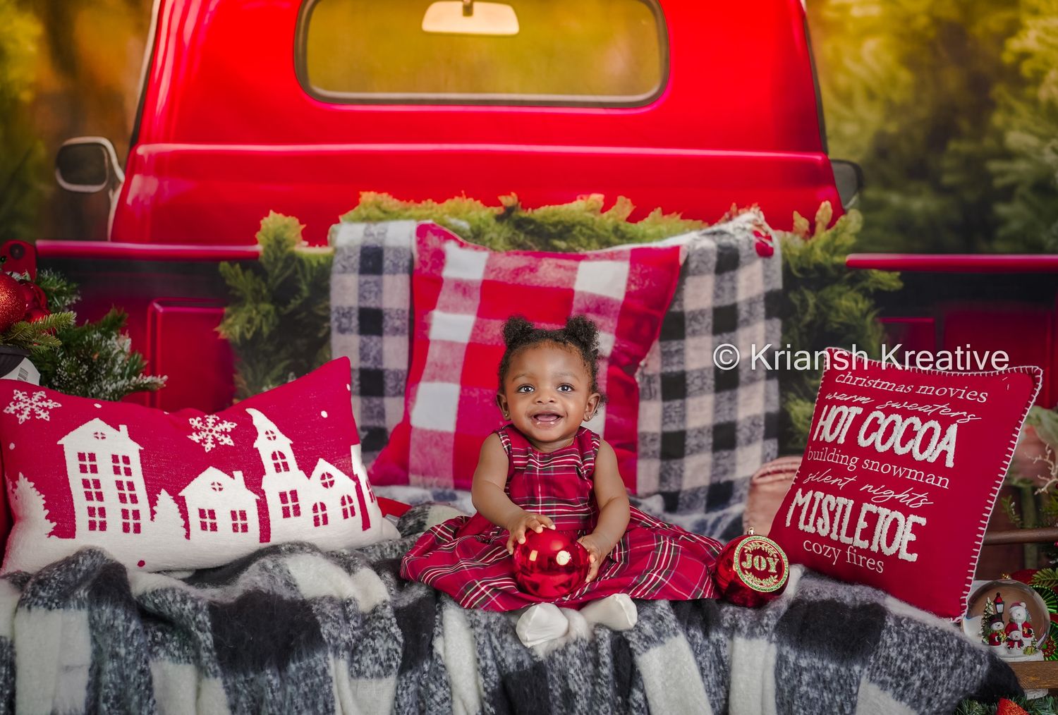 Festive Christmas setup with a red vintage truck backdrop, plaid blankets, decorative pillows and holiday decor elements.