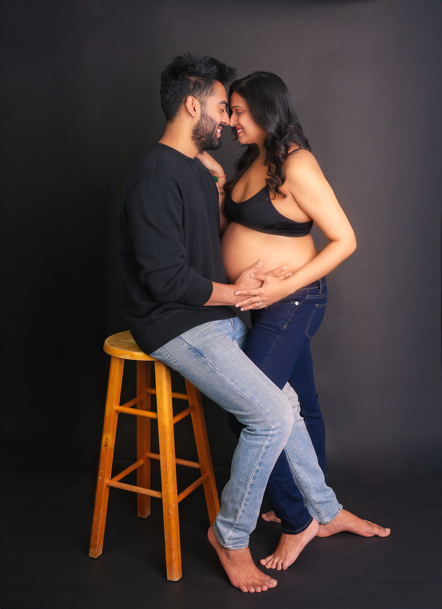 A romantic couple in casual clothing shares intimate moments posing together with a wooden stool against a dark background.