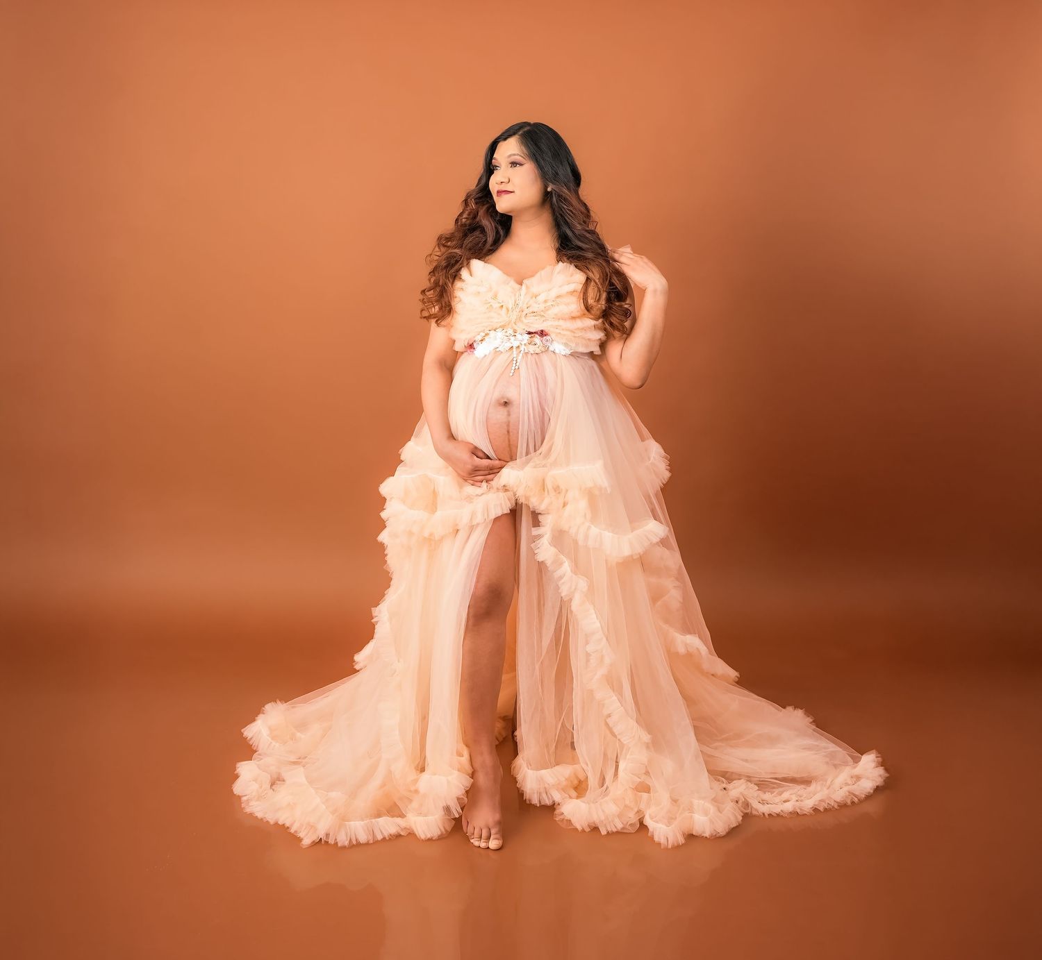 Elegant maternity photoshoot featuring flowing peach tulle gown against warm copper backdrop in studio lighting.