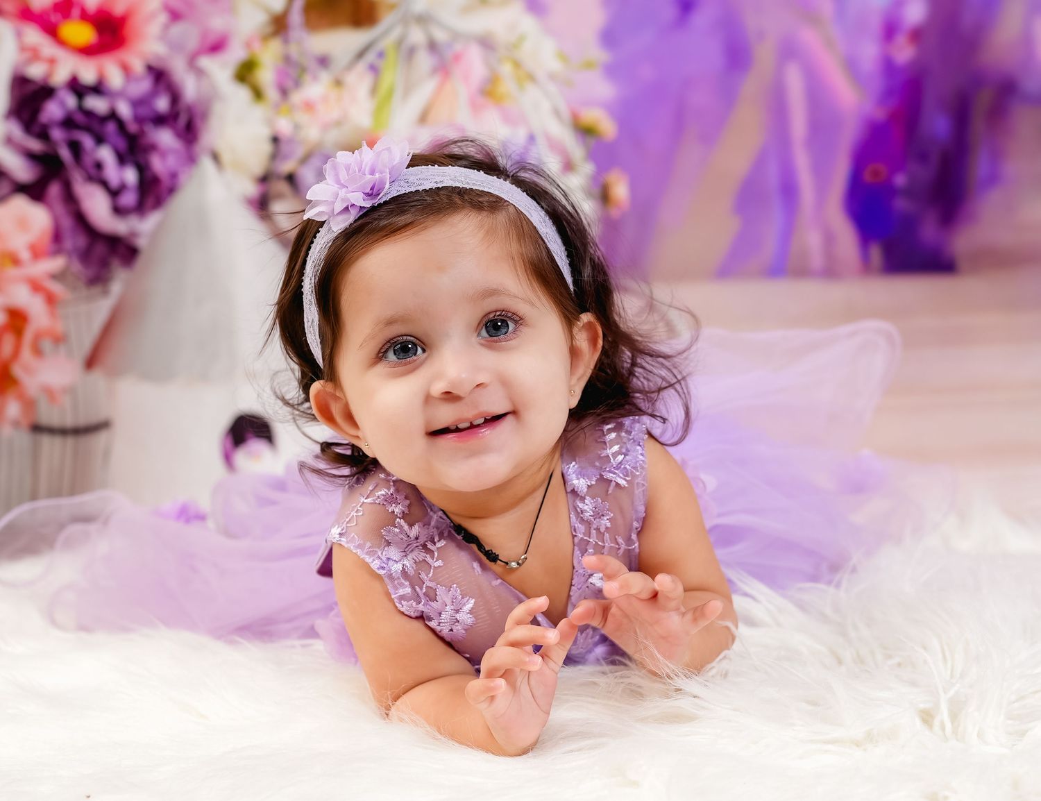 A small child in a purple dress lies on white tulle fabric against a colorful floral backdrop.