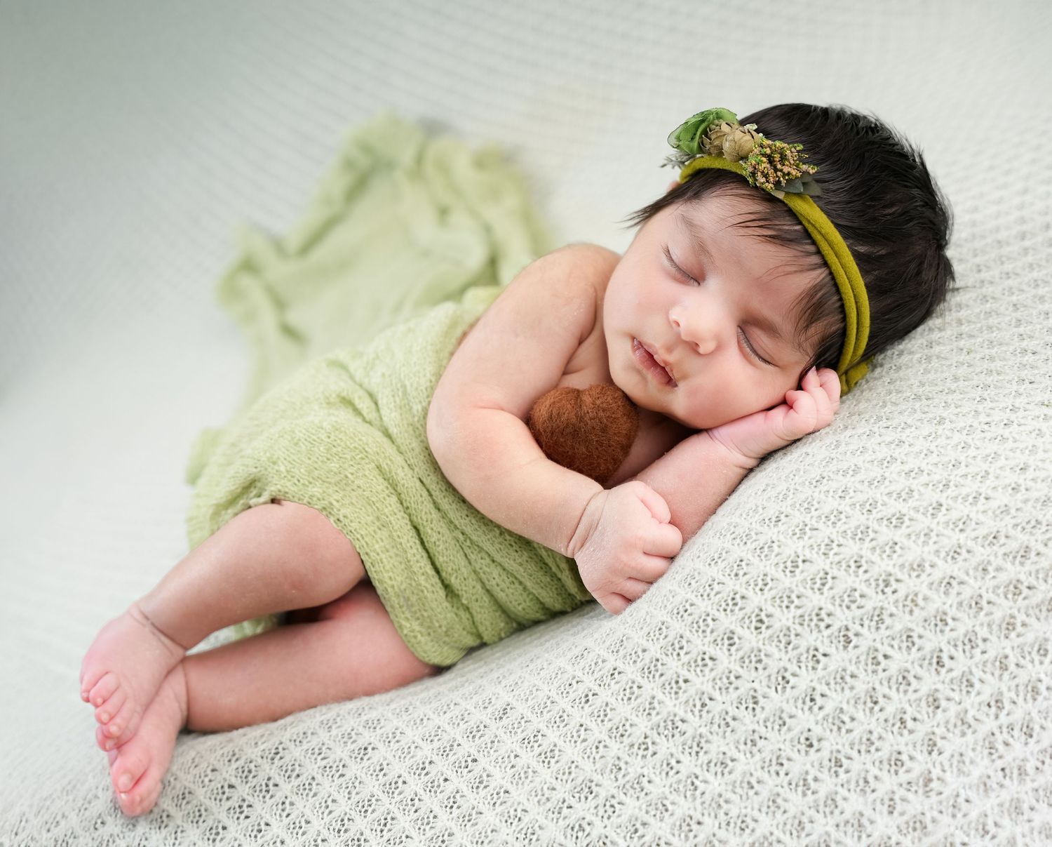 A sleeping newborn wrapped in soft green fabric lies on textured white fabric wearing a gold headband.
