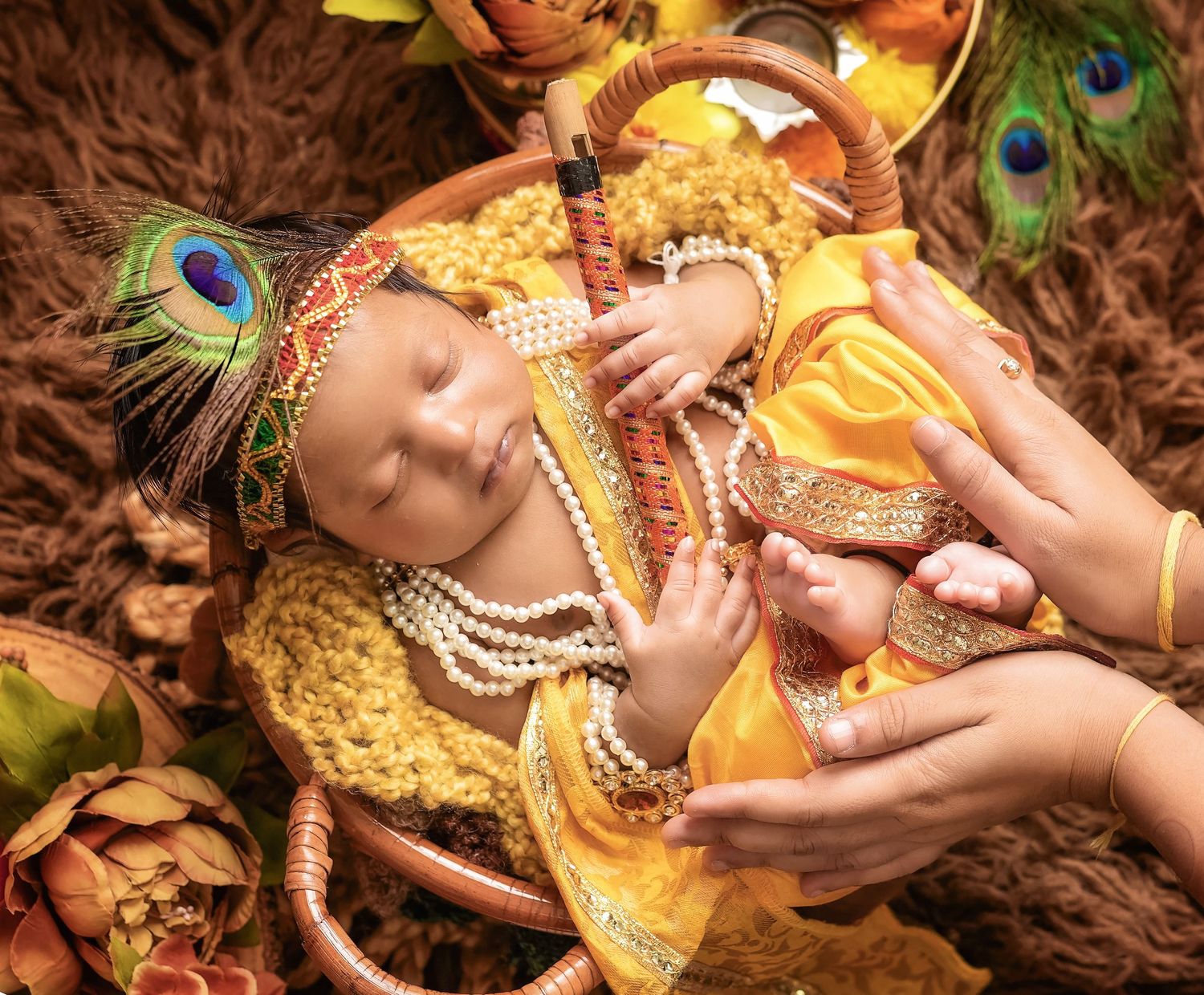A small baby dressed in traditional Krishna costume with peacock feathers lies peacefully on brown fabric with gentle hands nearby.