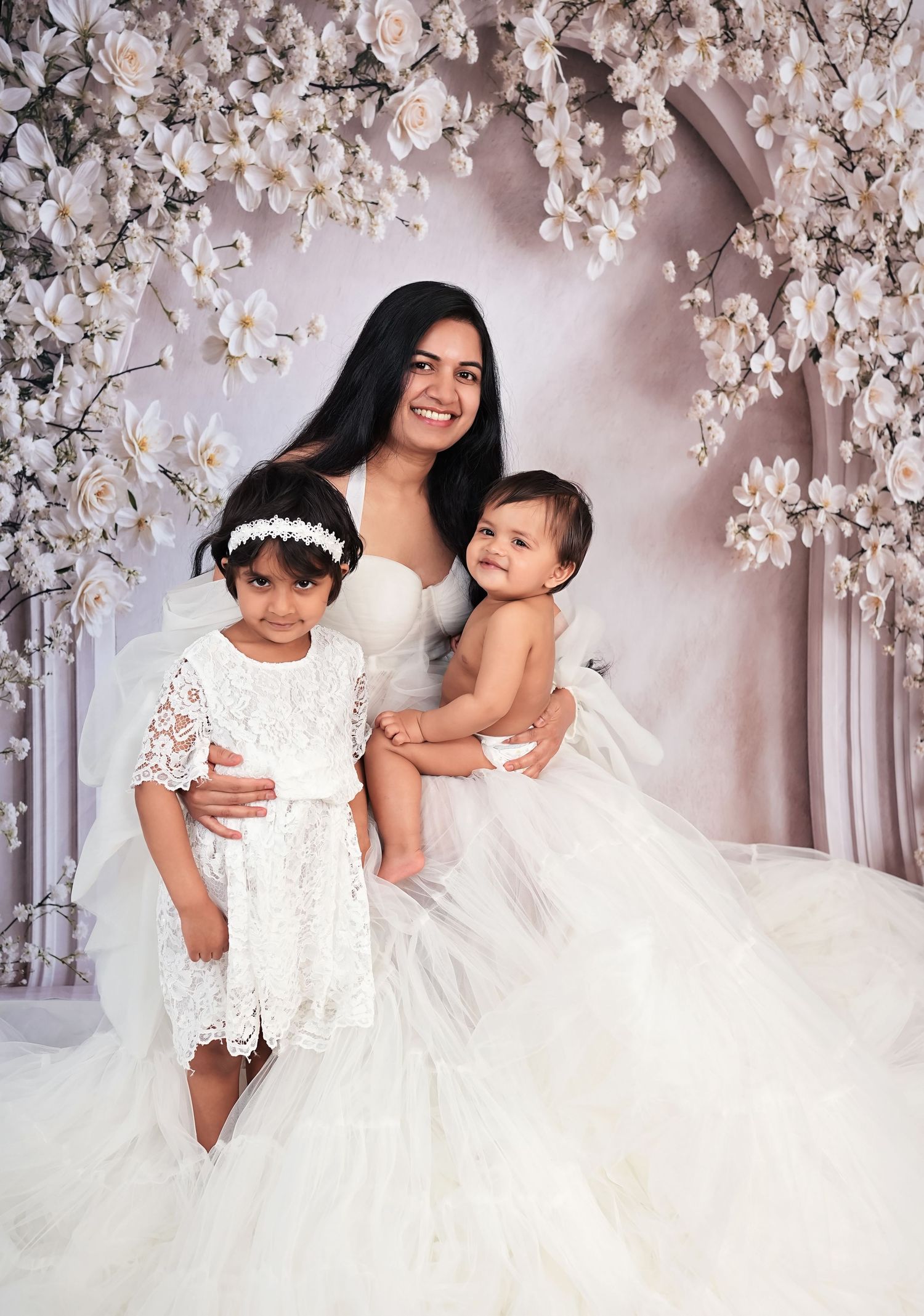 Woman in white bridal gown poses with two young girls in white dresses amid floral arch backdrop.