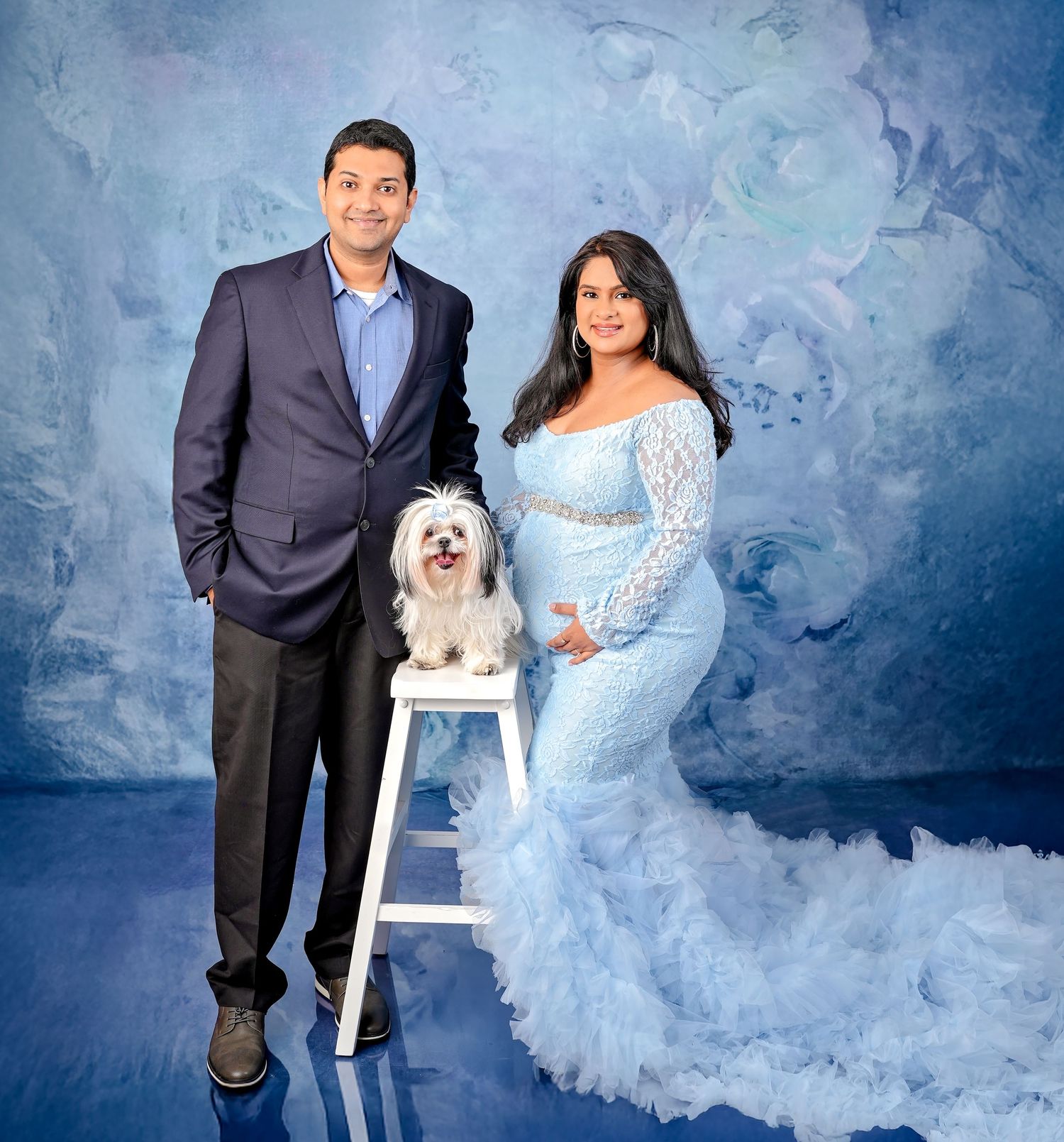 Elegant couple poses with small white dog on stool against blue textured backdrop in romantic portrait setting.