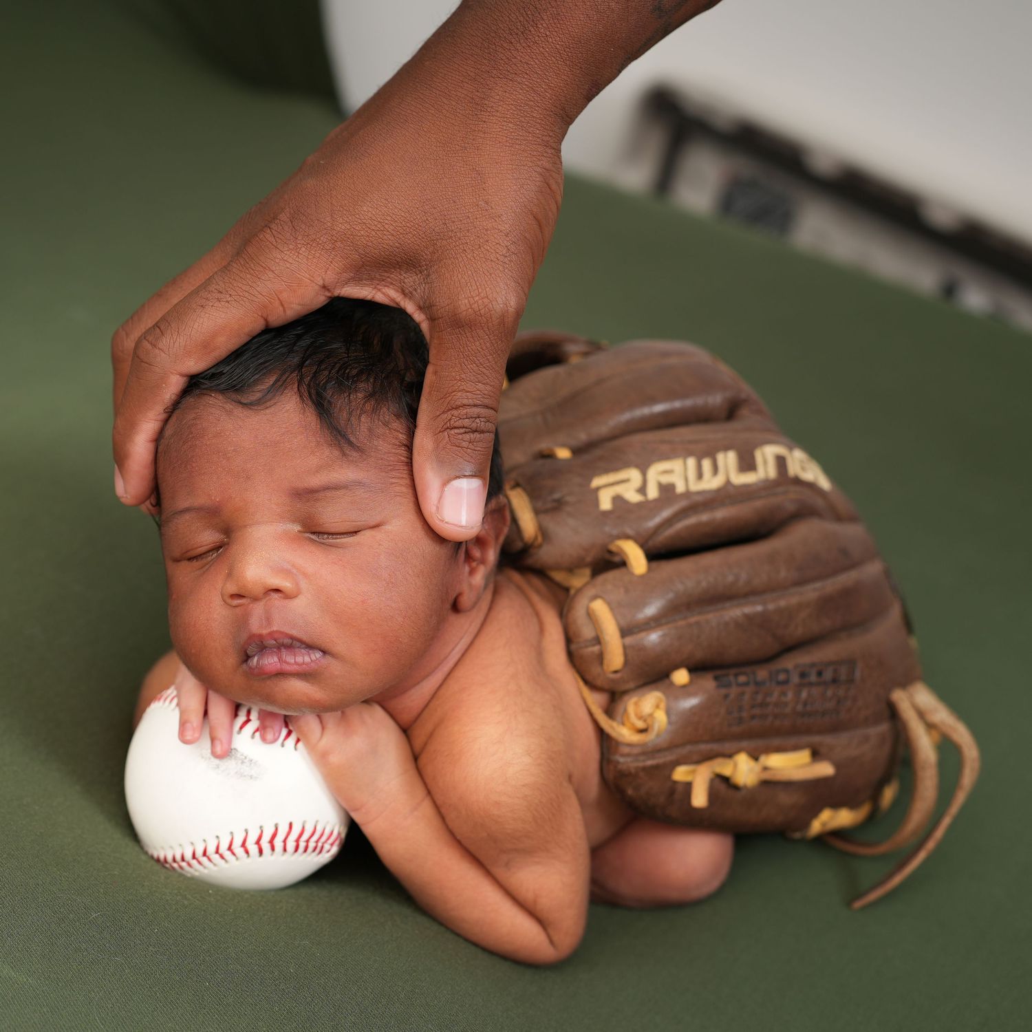 Sleeping newborn baby cradled in a Rawlings baseball glove resting on a baseball, with an adult hand gently touching its head.