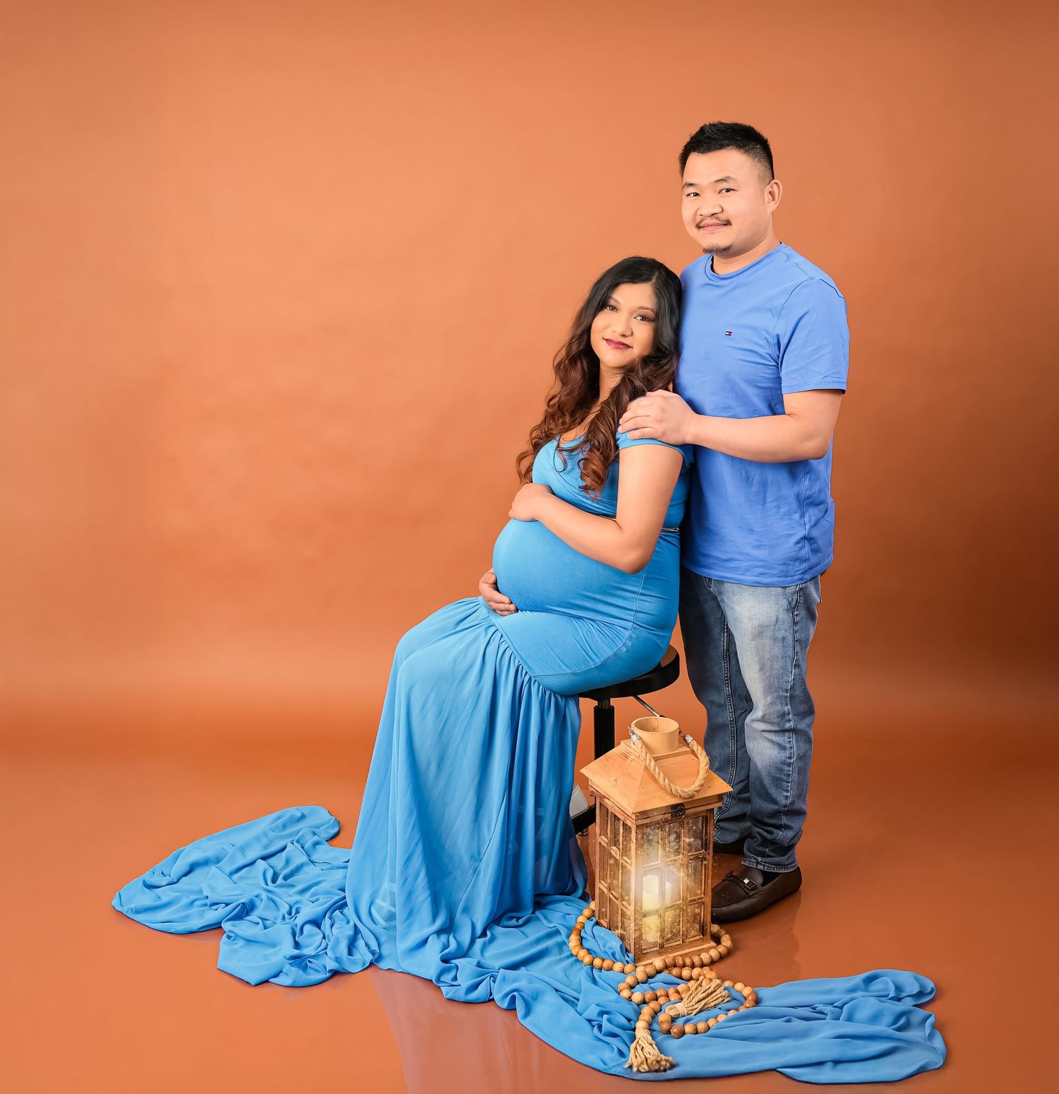 A couple in matching blue and denim outfits poses together with a woven basket in a portrait studio setting.