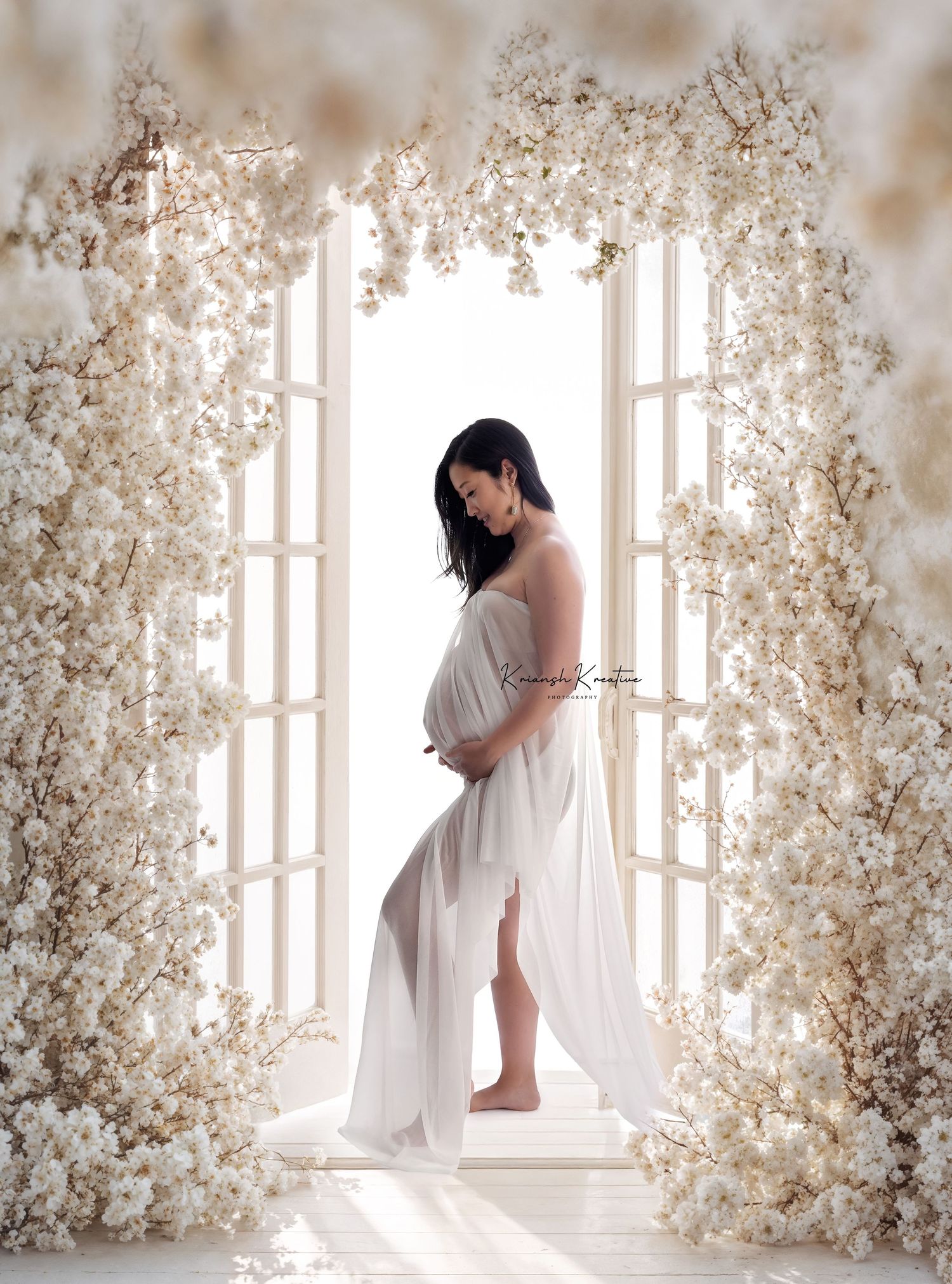 Ethereal maternity portrait in white flowing dress framed by delicate white floral archway in dreamy light.