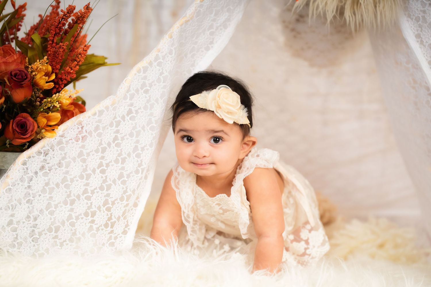 Baby wearing white floral headband sits among soft white textiles with orange flowers in background.