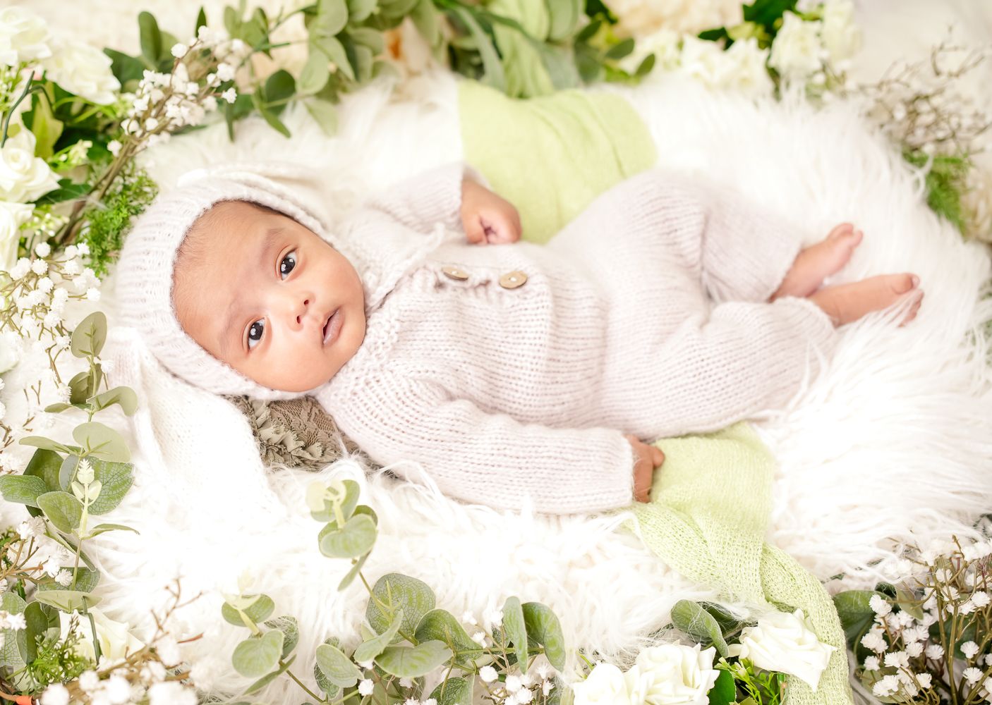 Baby in white knit outfit lying on soft white blanket surrounded by delicate greenery and floral decorations.