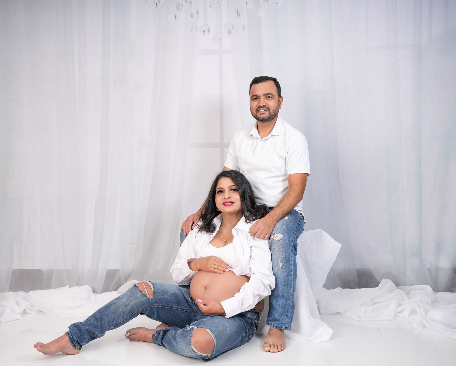 A pregnant woman in white shirt and jeans poses on white fabric with a person in casual attire behind her.