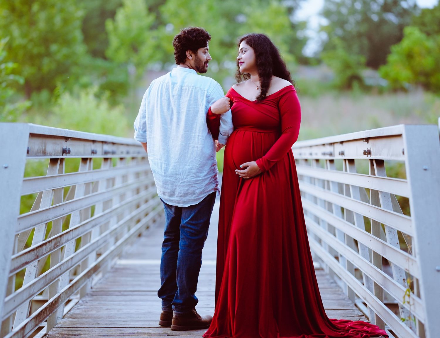 Couple stands on wooden boardwalk with expectant mother wearing flowing red maternity dress gazing at each other.