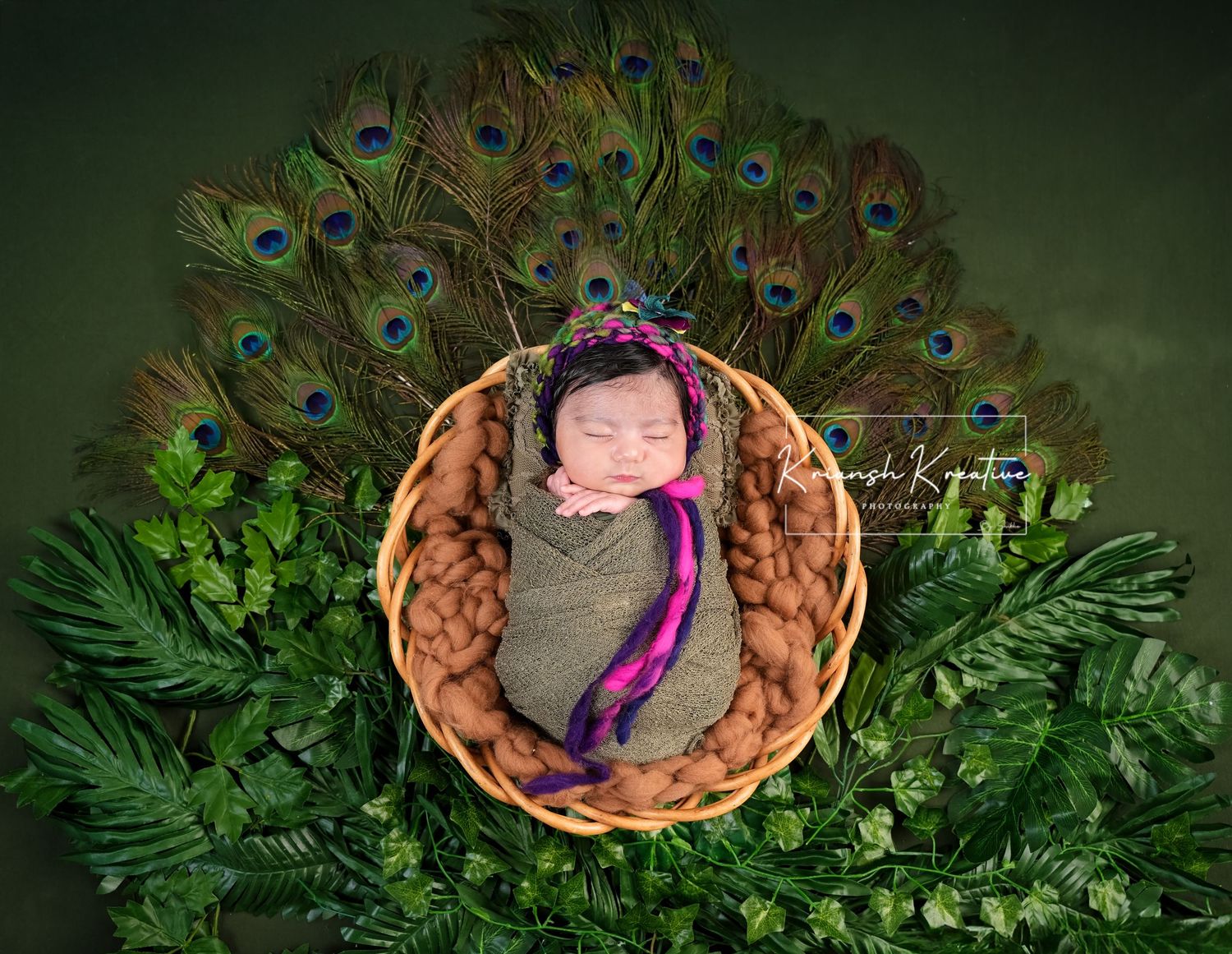 Sleeping newborn in woven basket surrounded by green ferns and vibrant peacock feathers on dark background.
