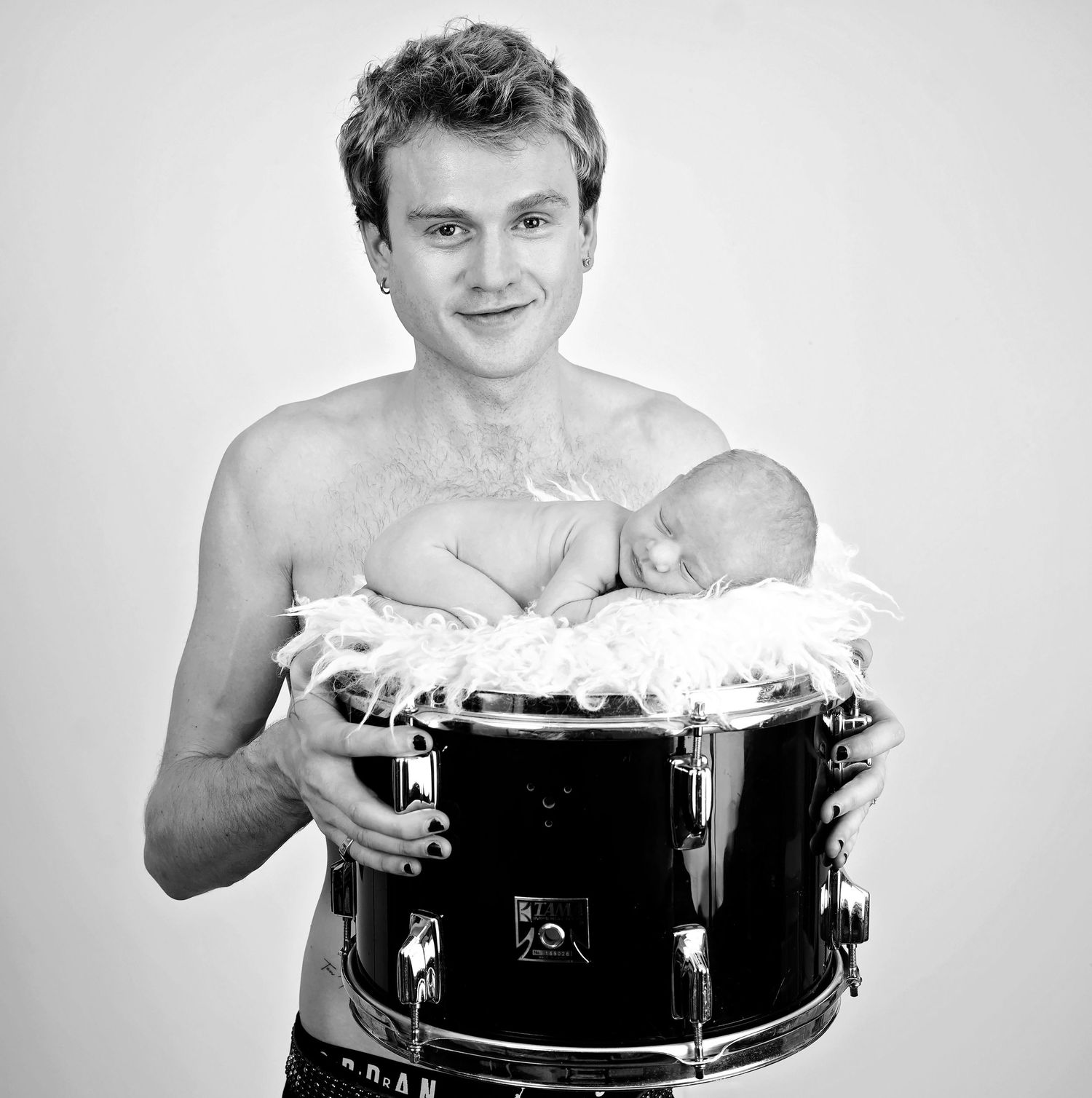 Black and white photo of a person posing shirtless with a drum while smiling at the camera.