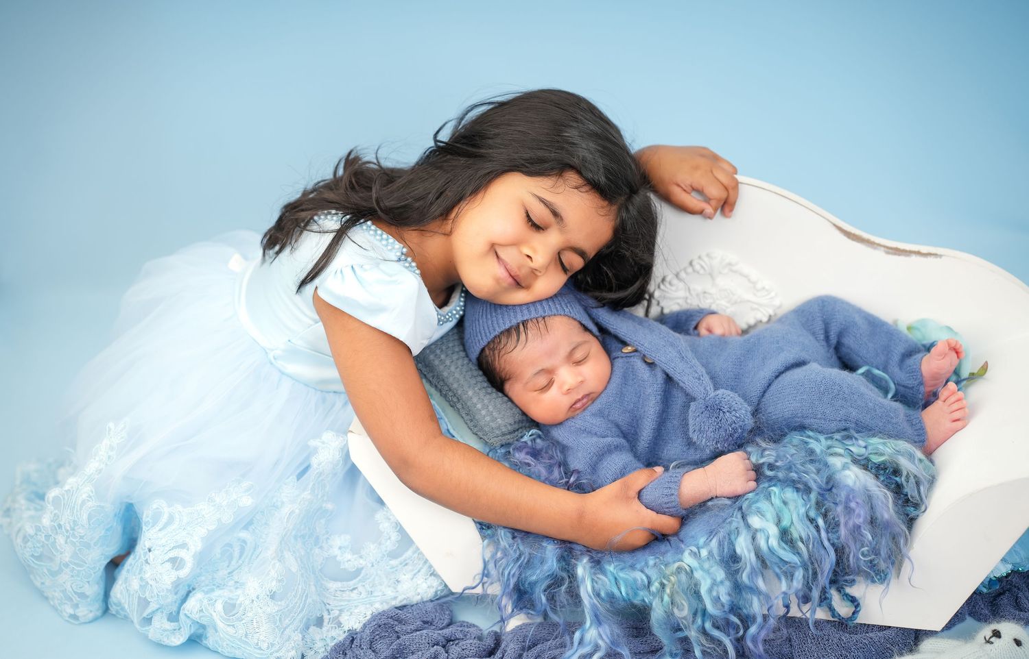 A young girl in a blue dress smiles while gently embracing her sleeping newborn sibling on a soft blue blanket.