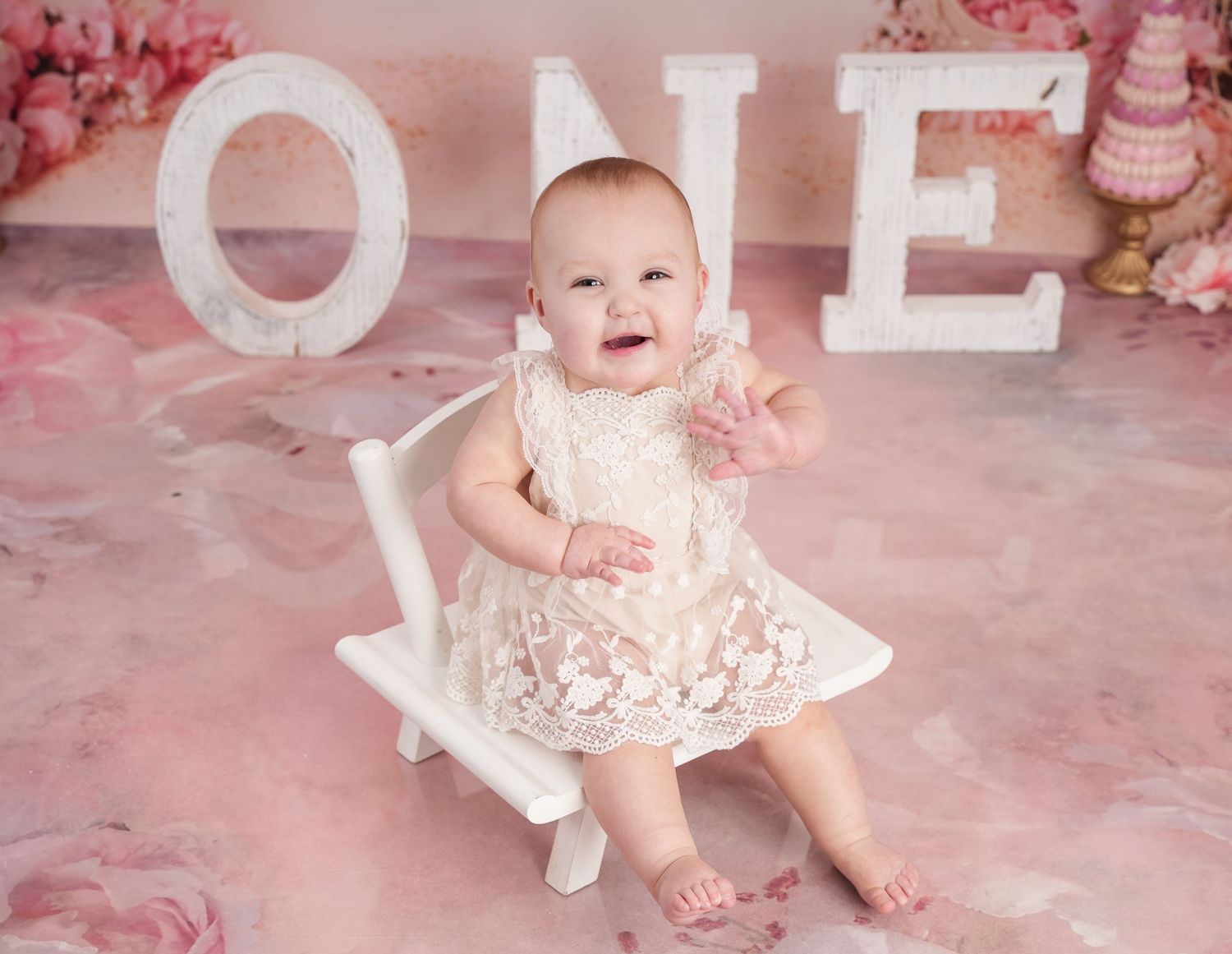Baby in a white lace dress sits on small chair against pink backdrop with 'ONE' letters for first birthday photoshoot.