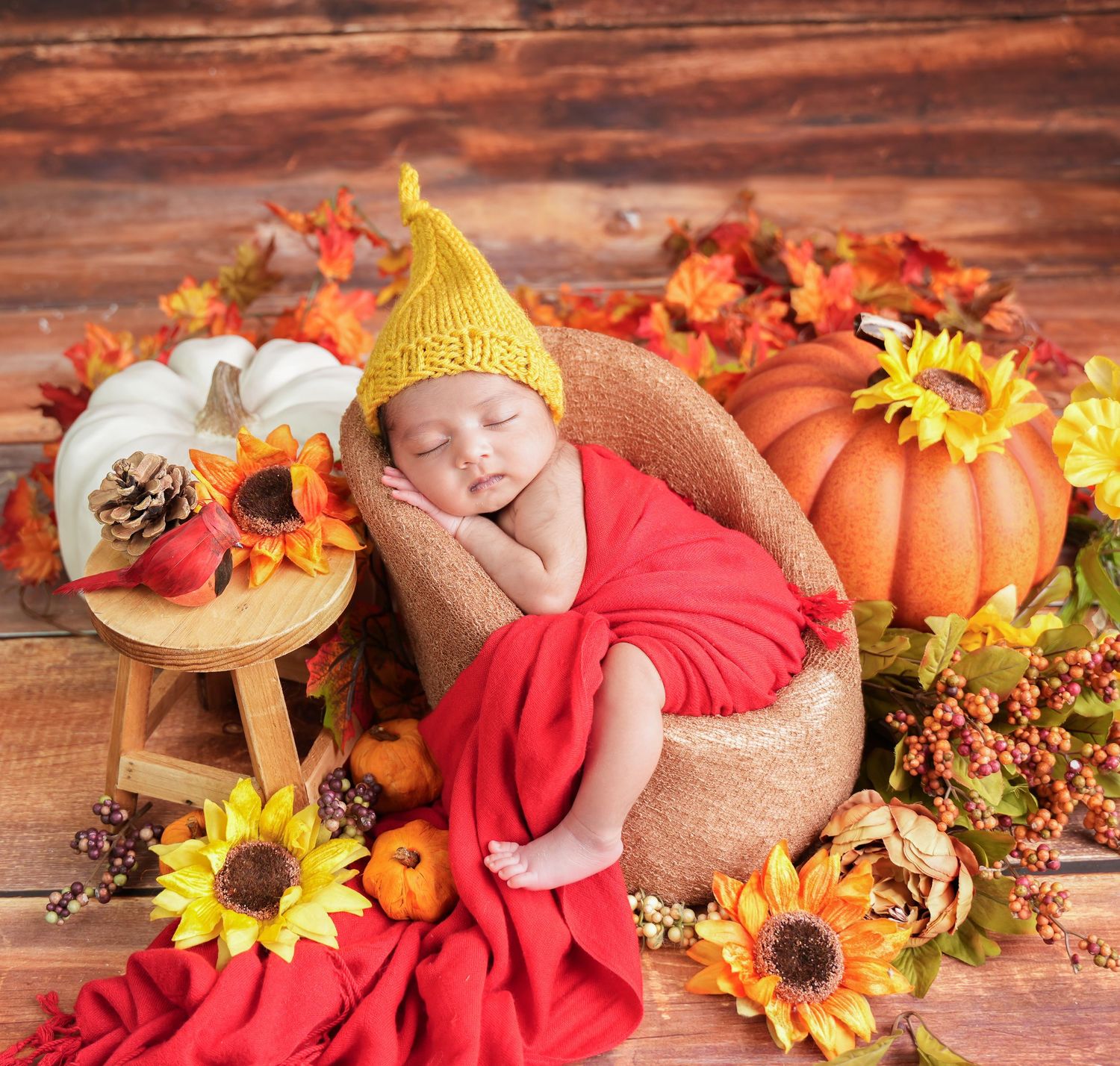 Newborn sleeping in autumn-themed setting with pumpkins, sunflowers, red fabric, and rustic wooden backdrop.