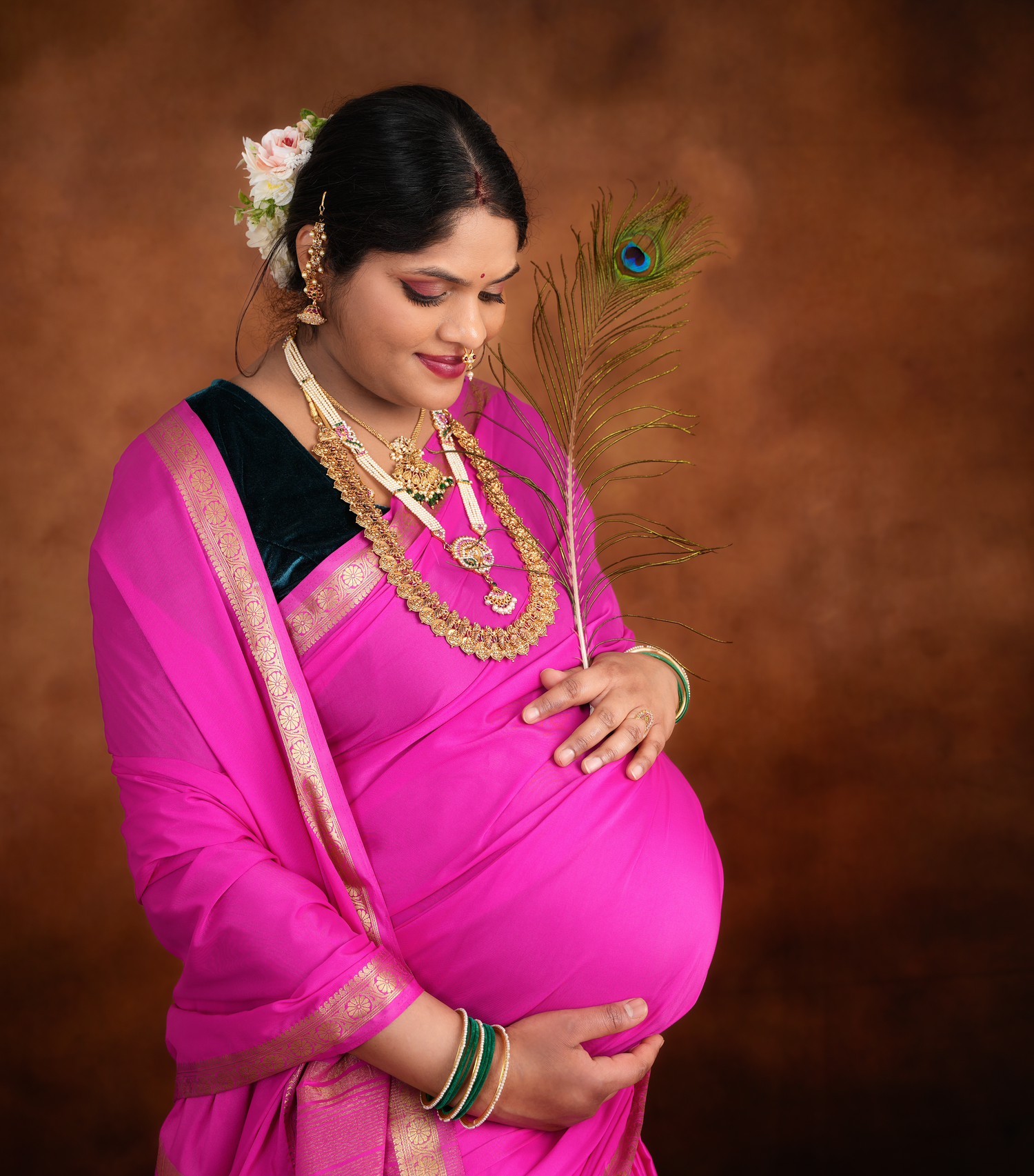 Maternity portrait of someone in a vibrant pink sari with gold jewelry and a peacock feather accent against brown backdrop.