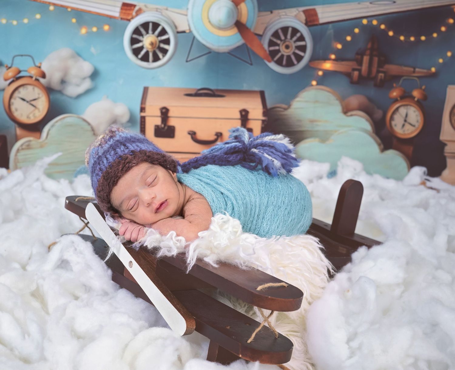 Sleeping newborn wrapped in blue swaddle lying on wooden airplane prop with cloud backdrop and travel theme decor.