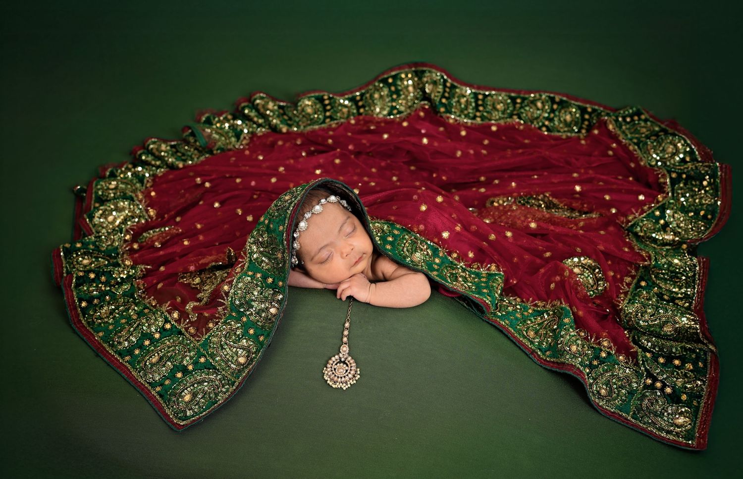 A newborn baby sleeps on a dark green blanket with a Christmas-themed red dress adorned with gold decorative accents.