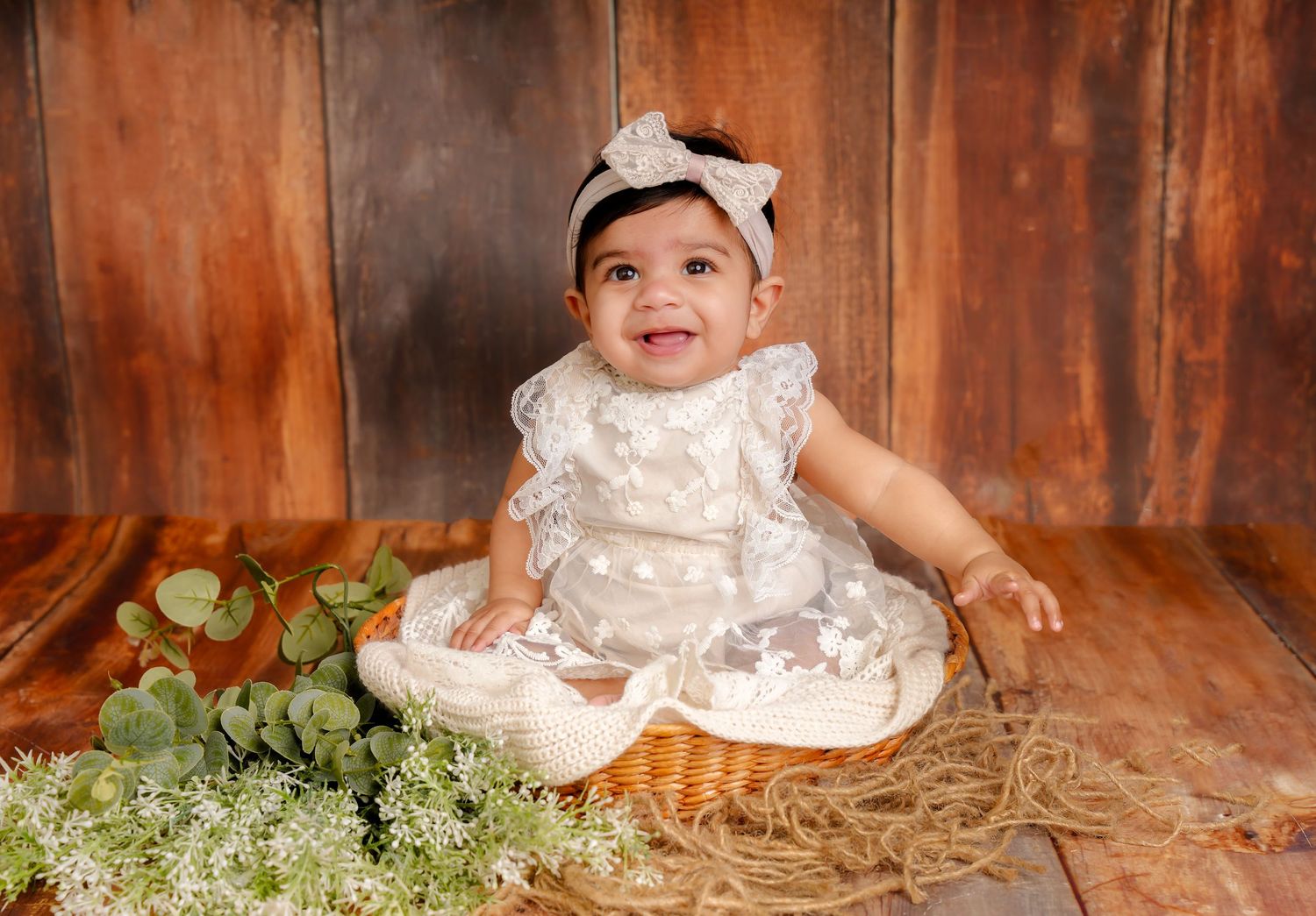 Sweet baby in white lace dress sitting on wooden floor with floral decorations for professional portrait session.