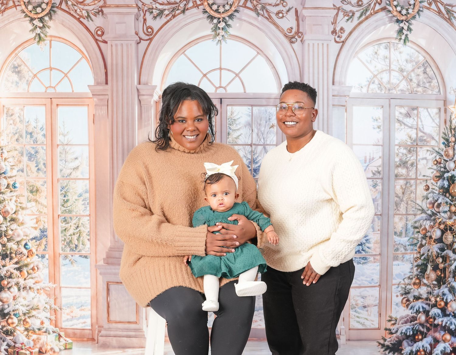 Family sits in front of decorated holiday backdrop with an ornate window and Christmas tree during winter photoshoot.