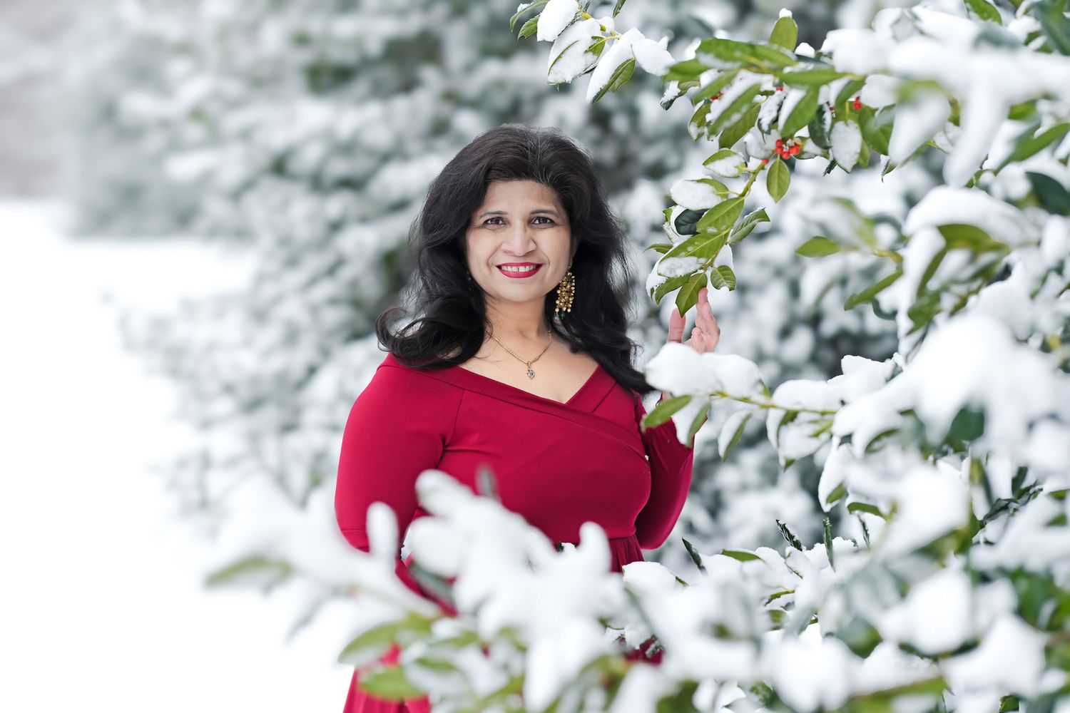 A person in a red sweater poses joyfully among snow-covered evergreen branches on a bright winter day.