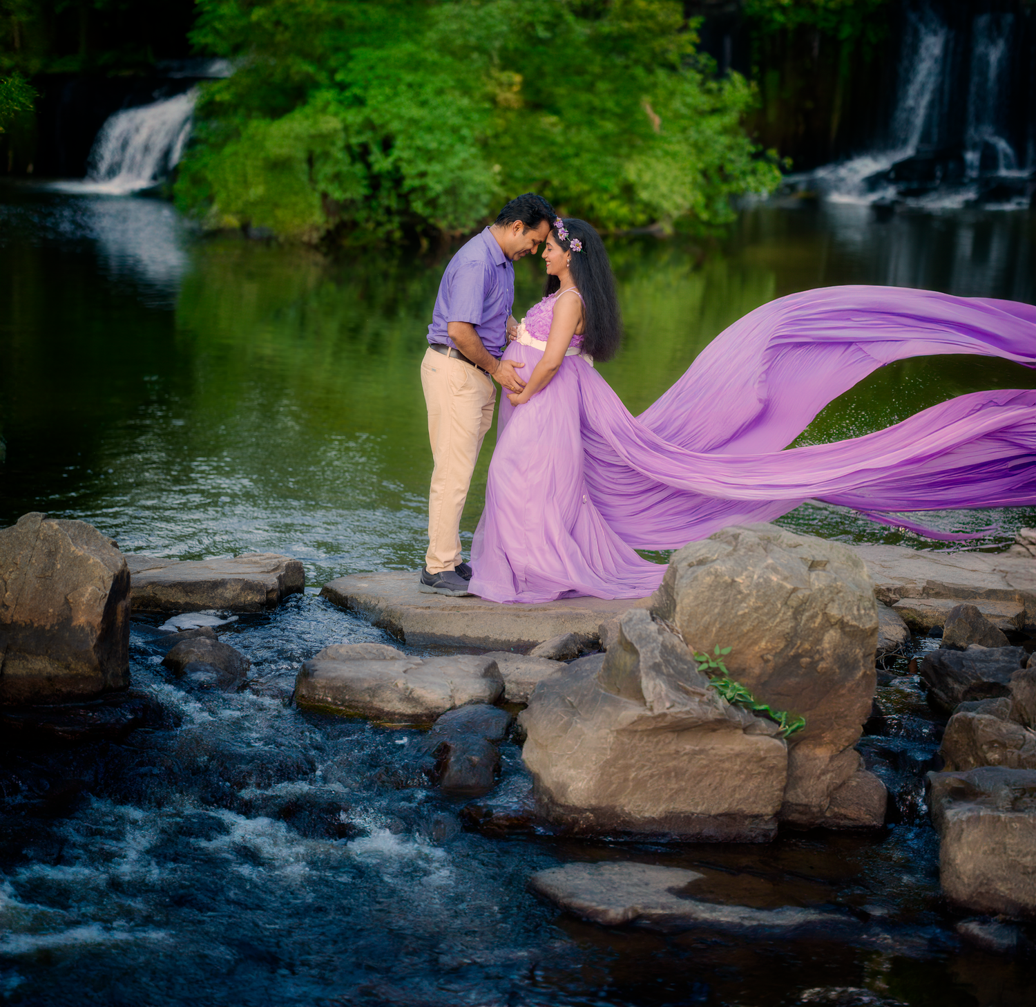 A couple embraces on rocky ledge by waterfall with flowing purple dress fabric creating dramatic movement in nature.
