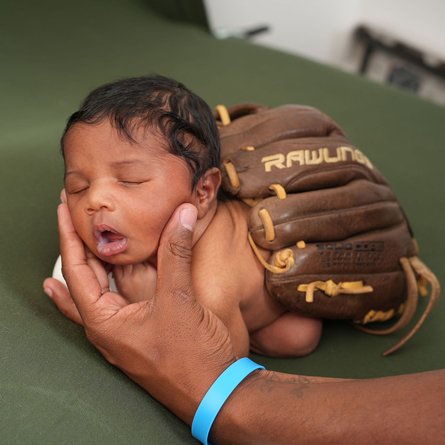 Newborn baby sleeping peacefully in a Rawlings baseball glove, cradled by adult hands wearing a blue wristband.