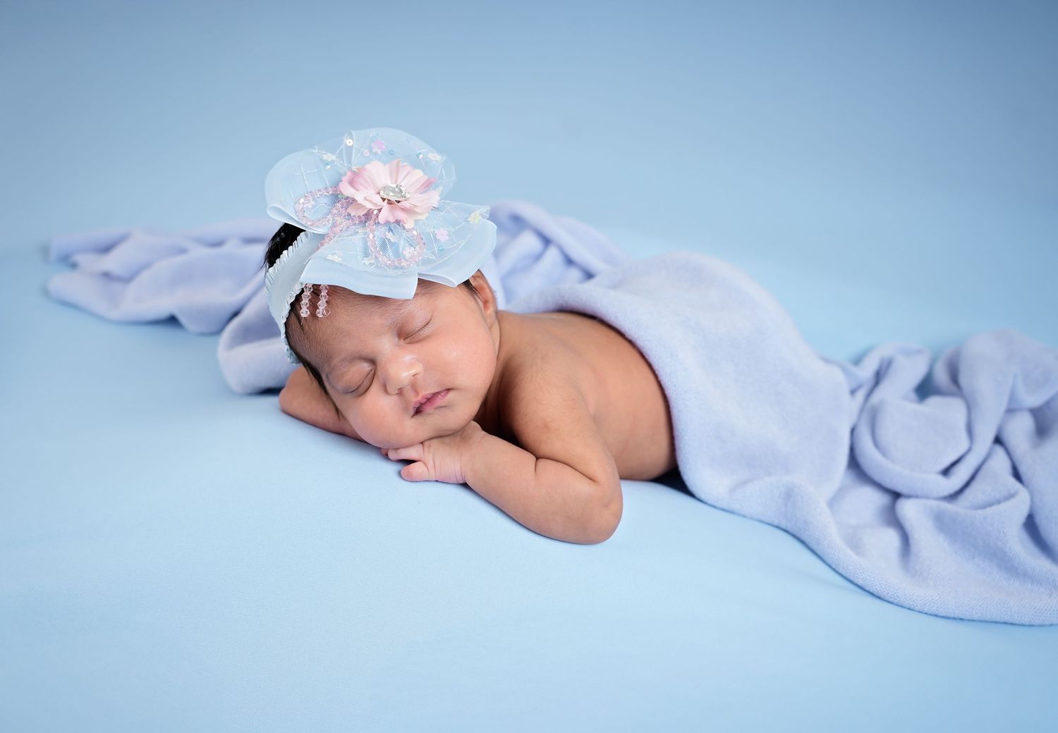 Sleeping newborn baby girl wrapped in a soft blue blanket wearing a white flower headband on a blue background.
