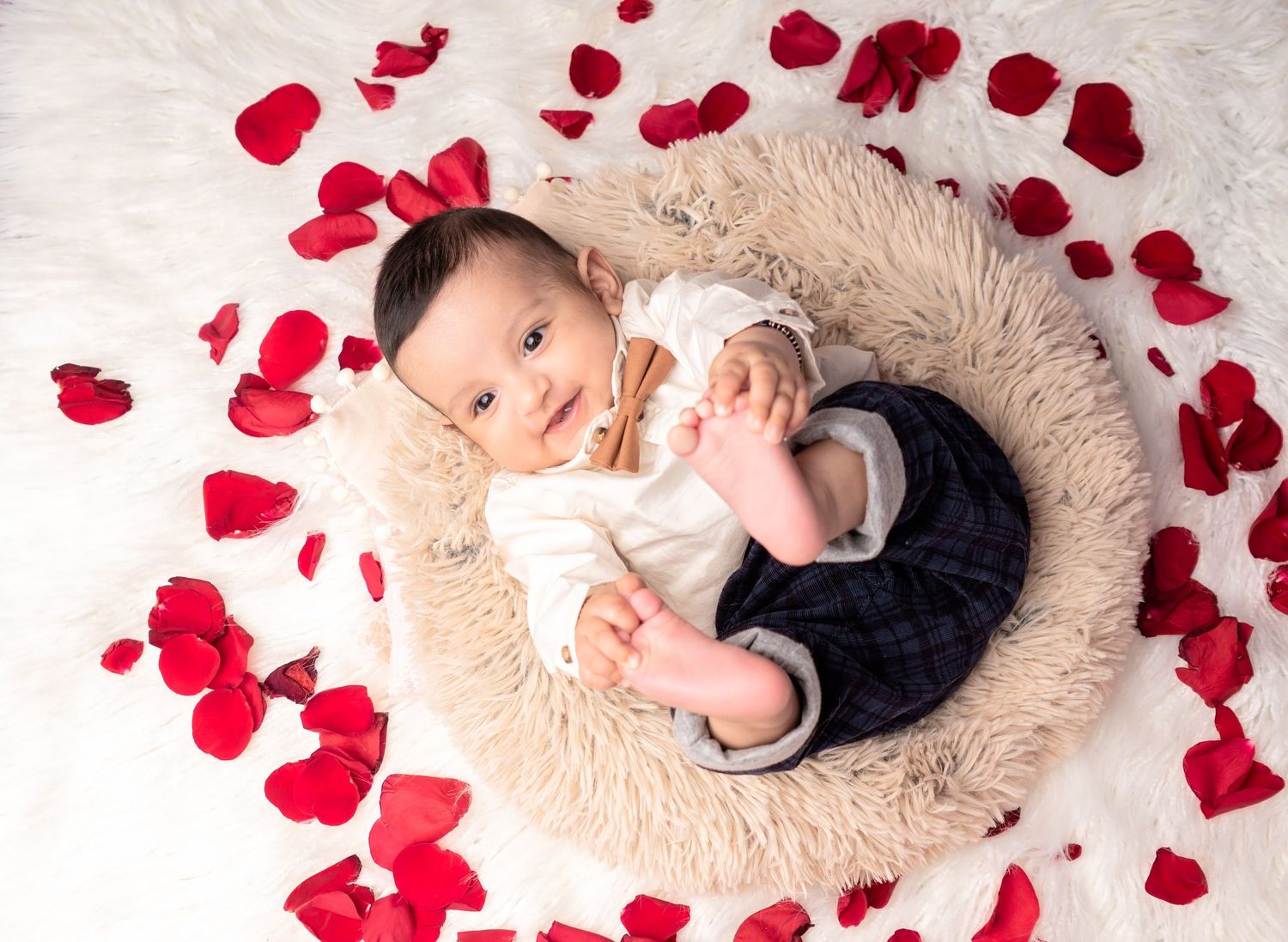 A baby lies in a cozy beige fur nest surrounded by scattered red rose petals in a studio photography setting.