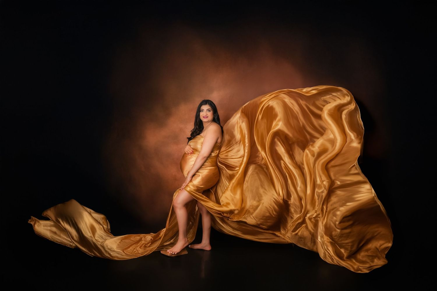Artistic maternity photo with flowing golden silk fabric against dark background creating dramatic effect.