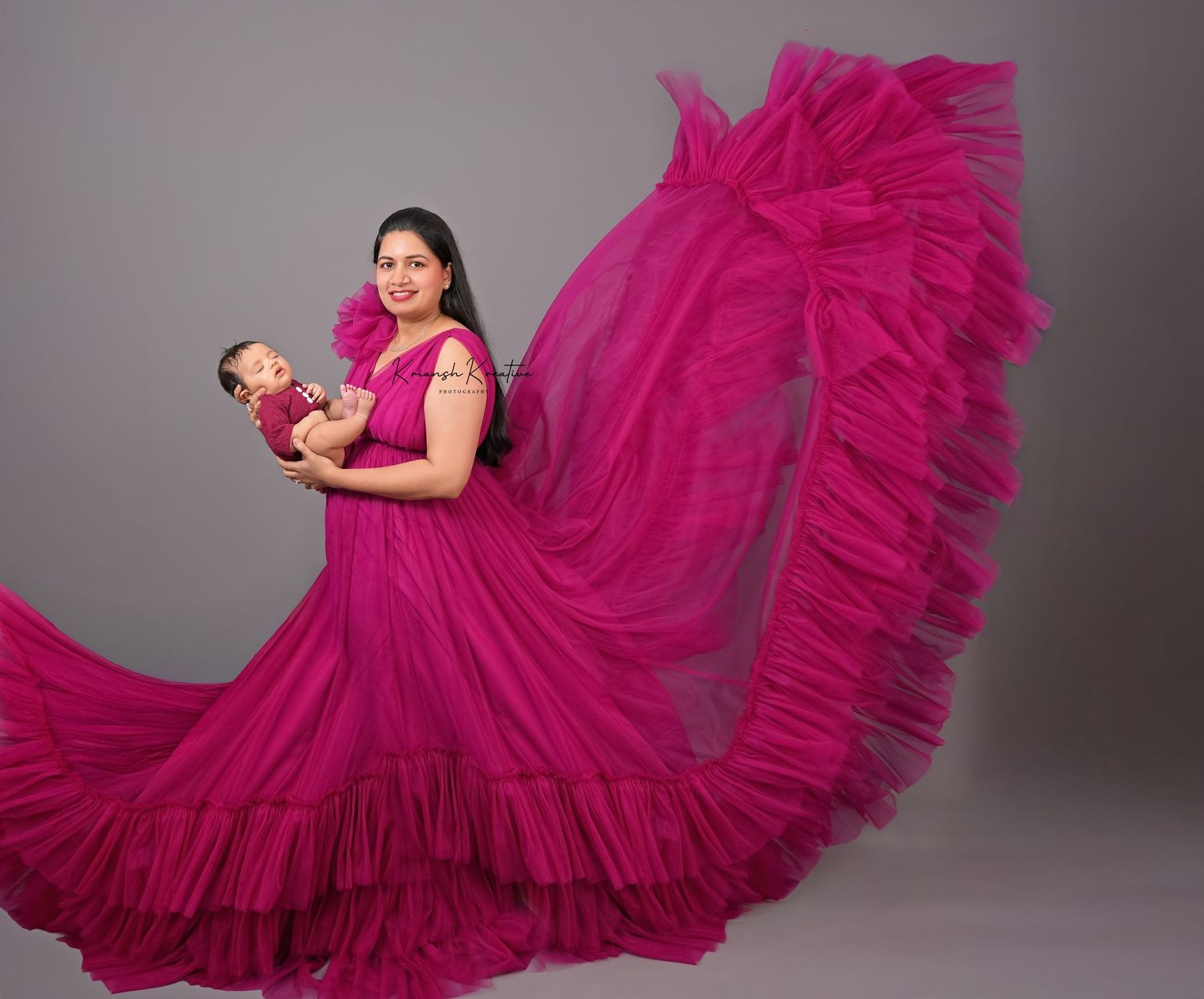 A magenta flowing ruffled gown with dramatic train sweeps across a gray studio backdrop during a maternity photo session.
