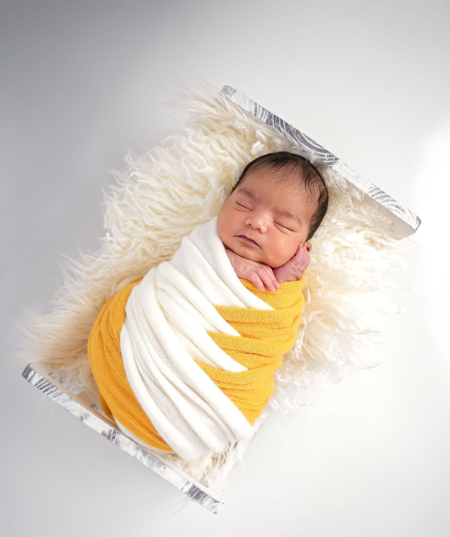 A newborn wearing a yellow wrap rests peacefully on white fur bedding in a soft studio light setup.