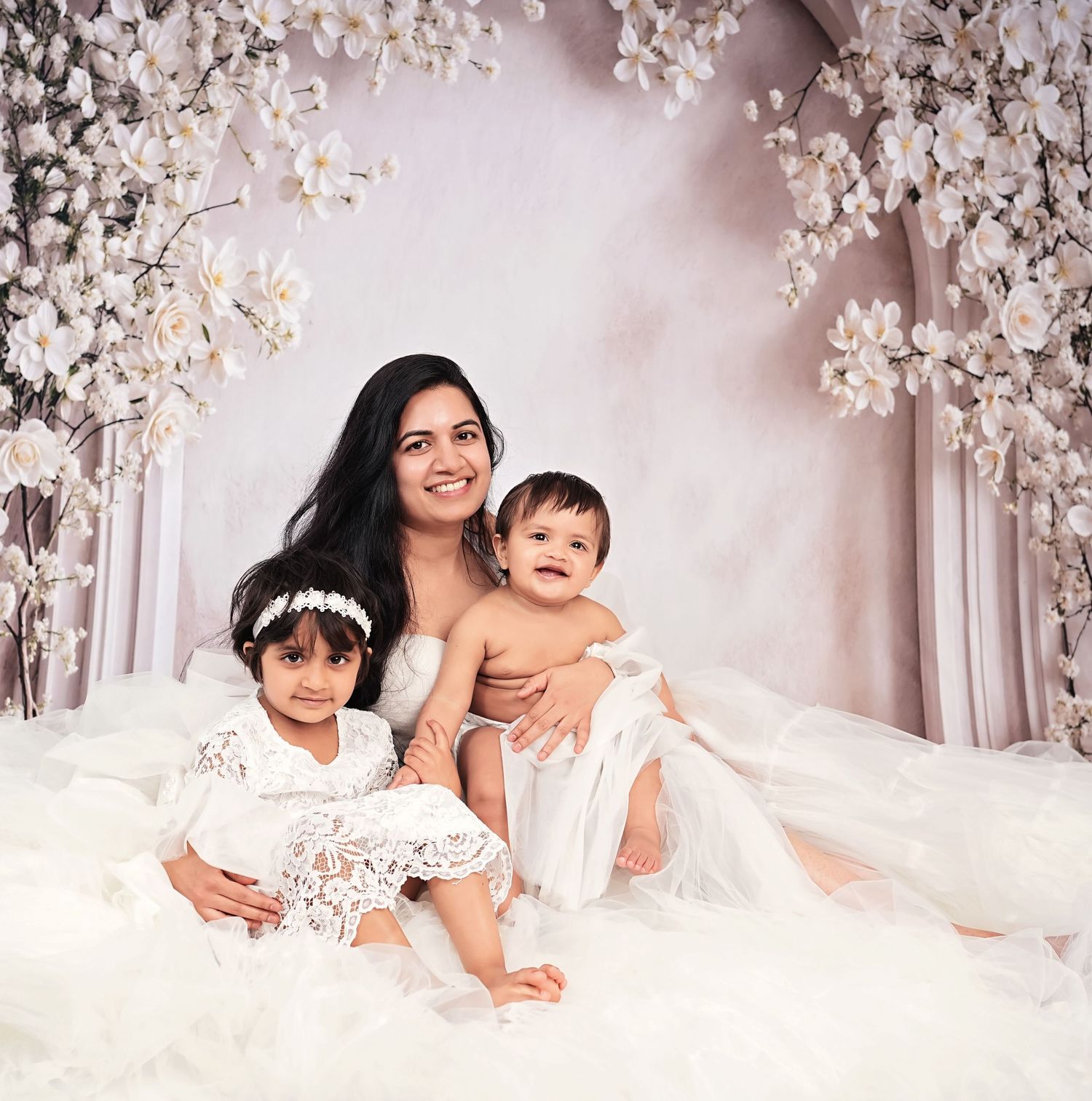 A mother in white dress poses with two young children against a floral backdrop in an elegant portrait.
