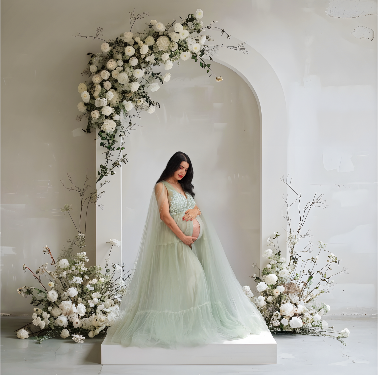 A romantic maternity portrait with a flowing mint green tulle gown surrounded by white floral arrangements against a white wall.