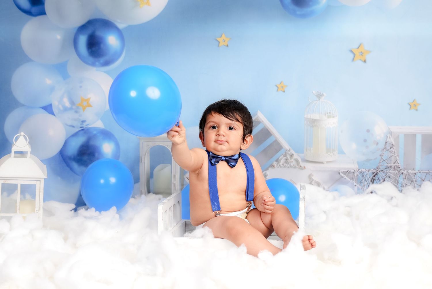 Baby wearing blue bow tie sits among blue balloons and stars in dreamy white studio setting with lanterns.