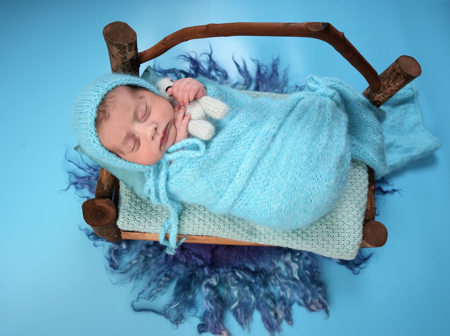 A sleeping newborn baby swaddled in light blue blanket rests on a small wooden bed with a blue background.