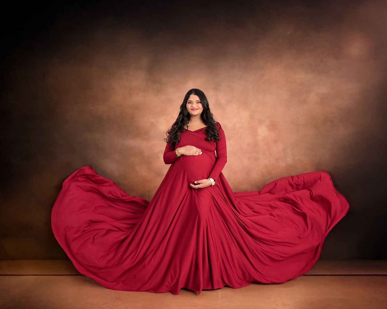 A flowing red maternity gown dress billows dramatically against a dark brown studio backdrop.