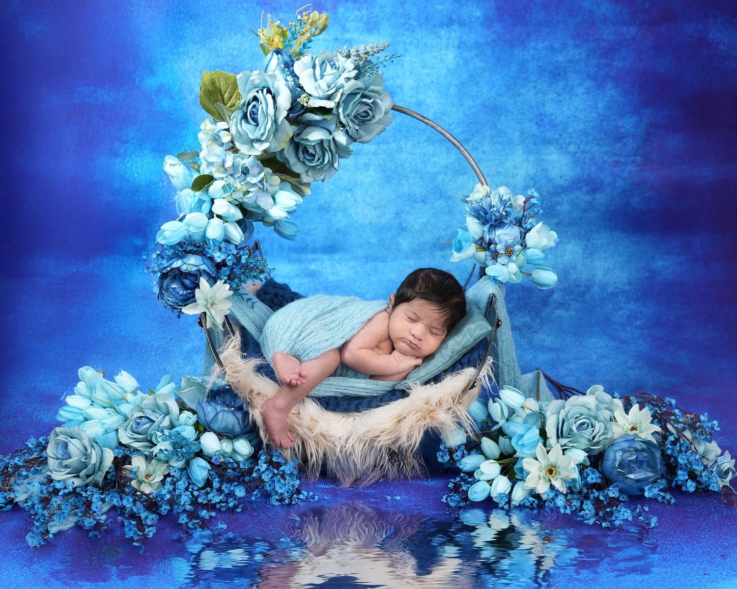 A sleeping baby rests in a crescent moon prop surrounded by blue flowers against a dreamy blue backdrop.