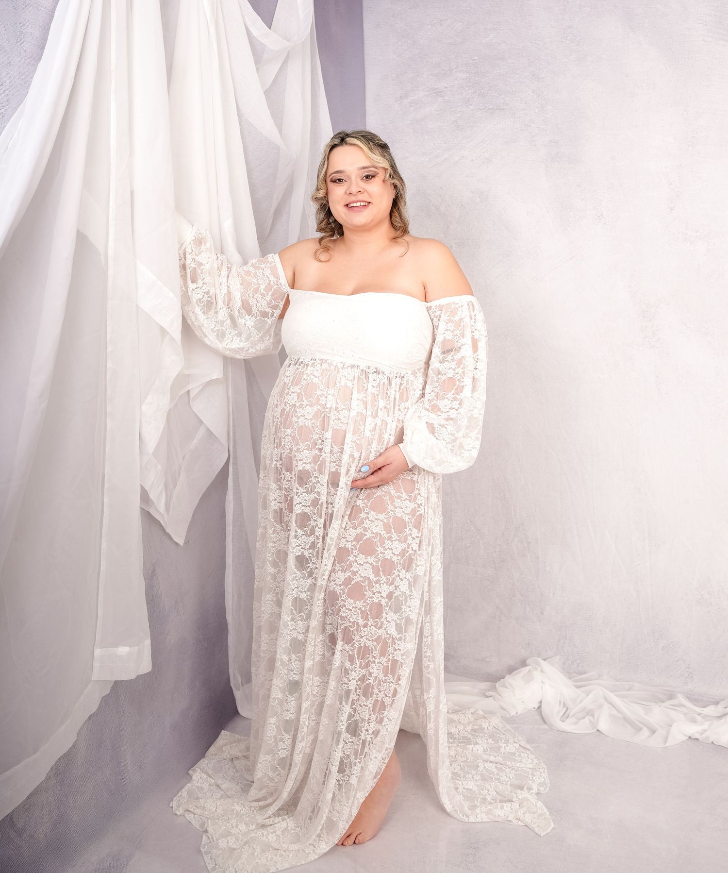 Ethereal white lace maternity dress displayed in a romantic studio setting with flowing sheer fabric background.