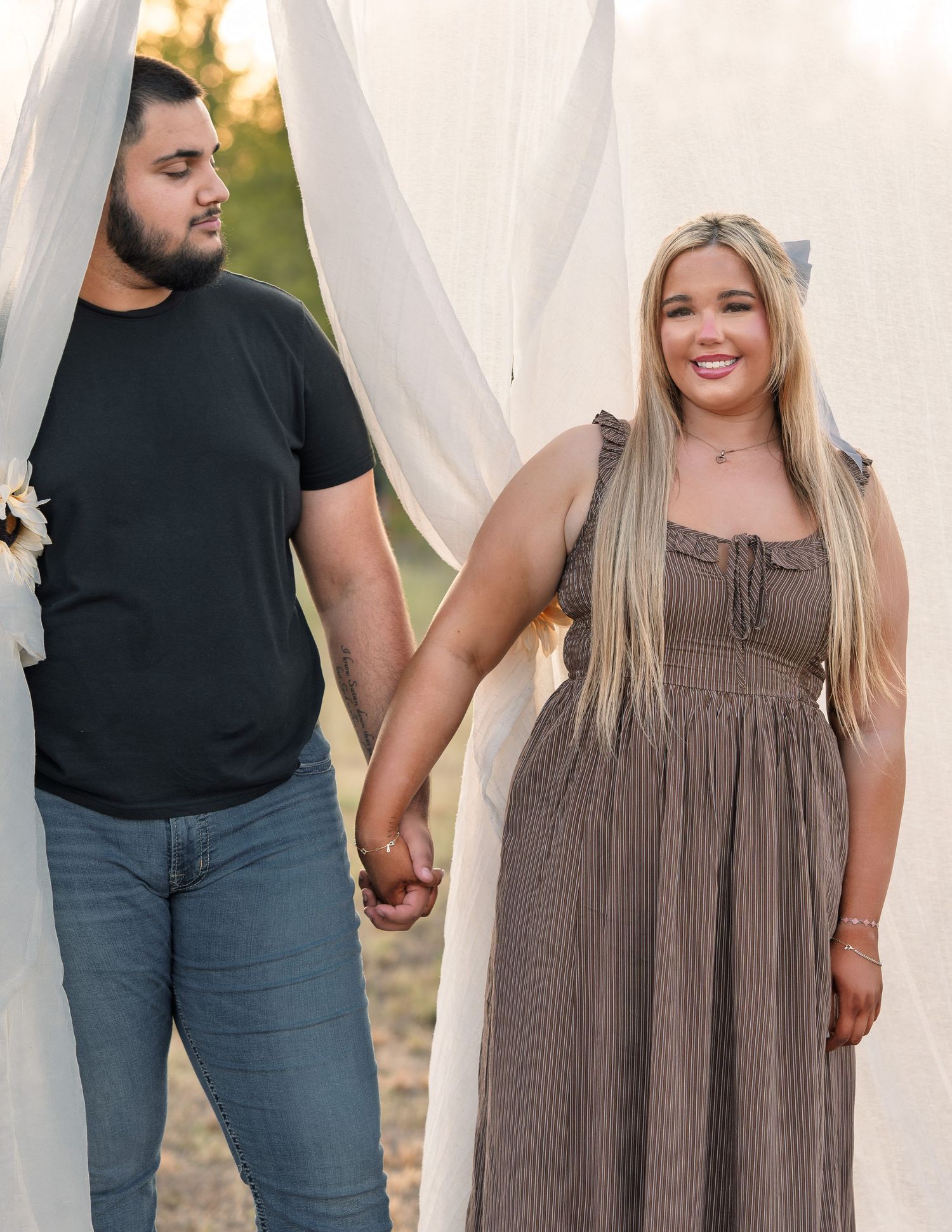 Couple holding hands smiling between white draping curtains during golden hour engagement photos.