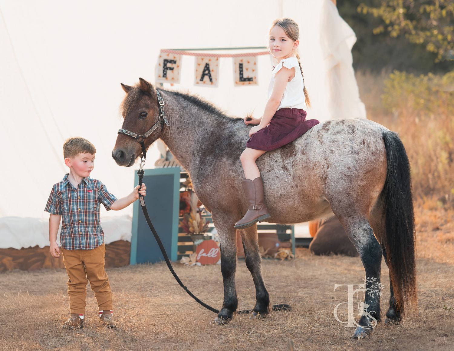 Young boy leading a pony with a girl sitting on it near a fall-themed backdrop.