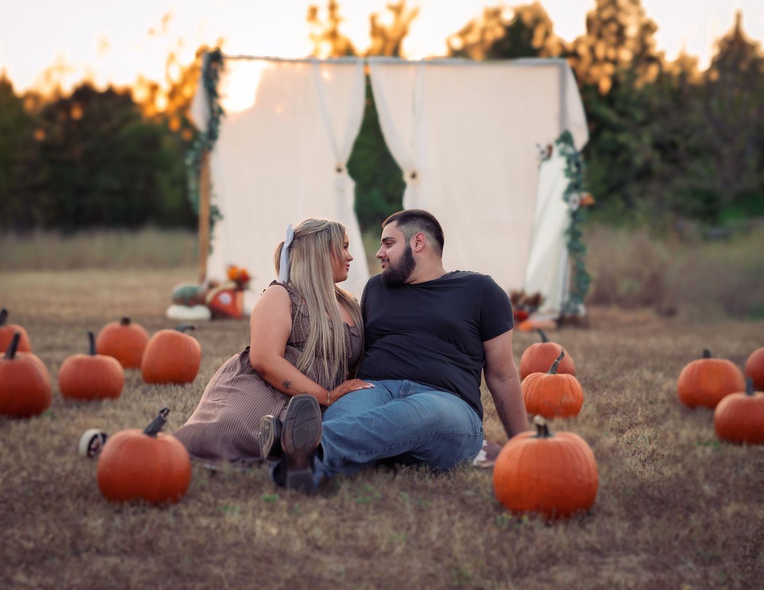 Couple sitting together in pumpkin patch at golden hour with white arch backdrop behind them.