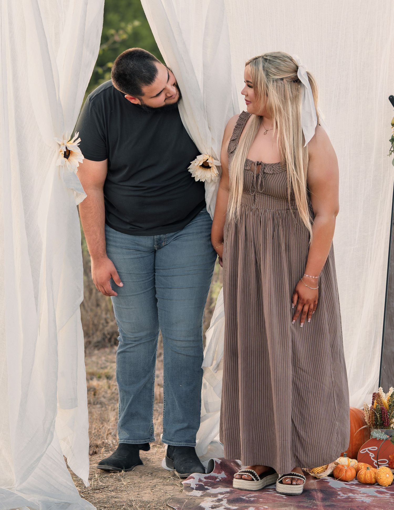 Couple sharing a tender moment between white fabric drapes with sunflower decorations at sunset.