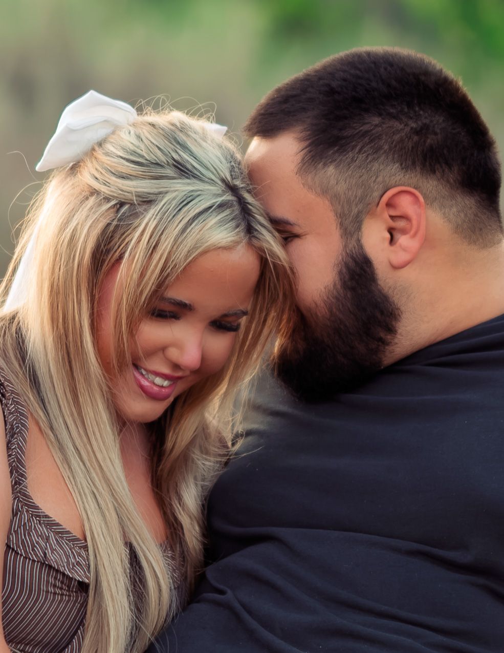 Close romantic moment of smiling blonde woman and bearded man touching foreheads outdoors.