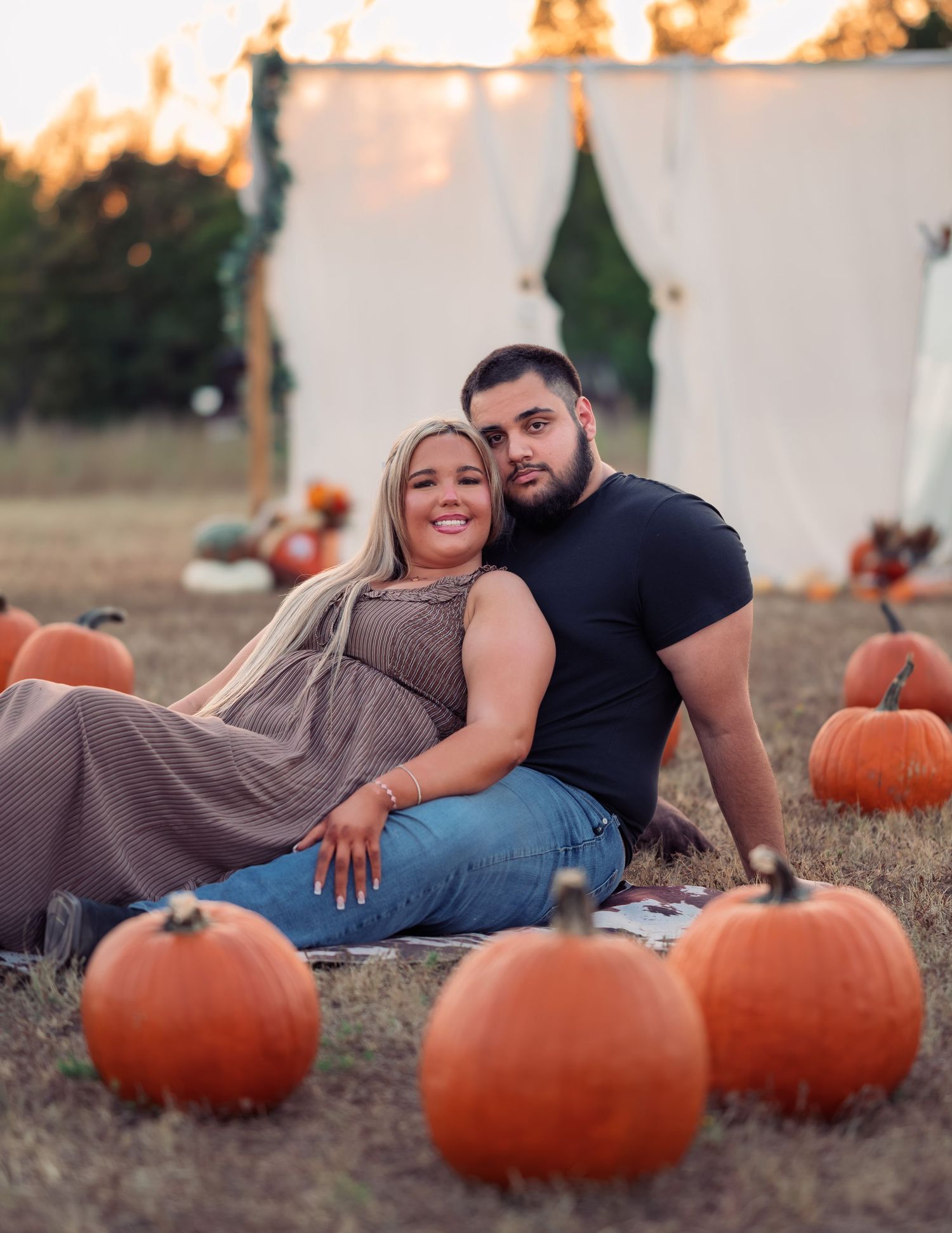 Couple smiling together on blanket surrounded by pumpkins with white arch backdrop at sunset.