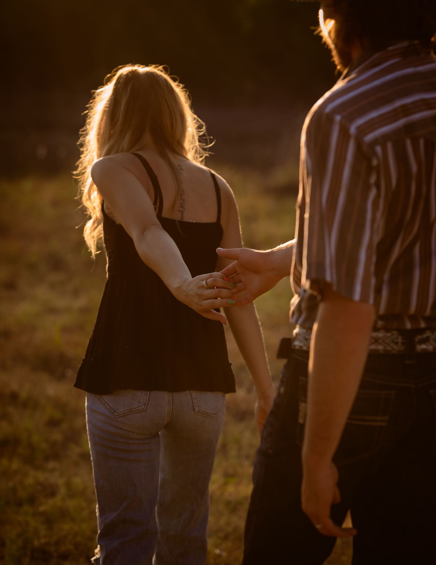 Couple holding hands walking through a golden sunlit field during sunset engagement shoot.